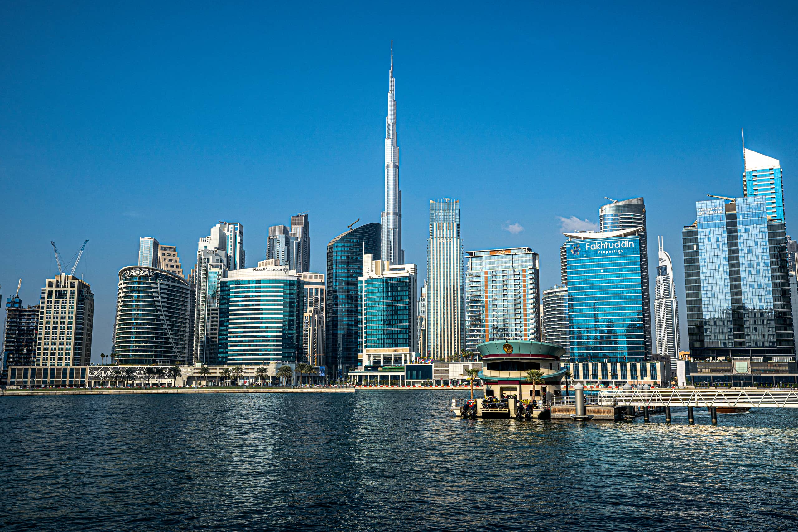 Wide daylight view across a body of water toward a dense city skyline, dominated by a towering skyscraper at the center under a clear blue sky.