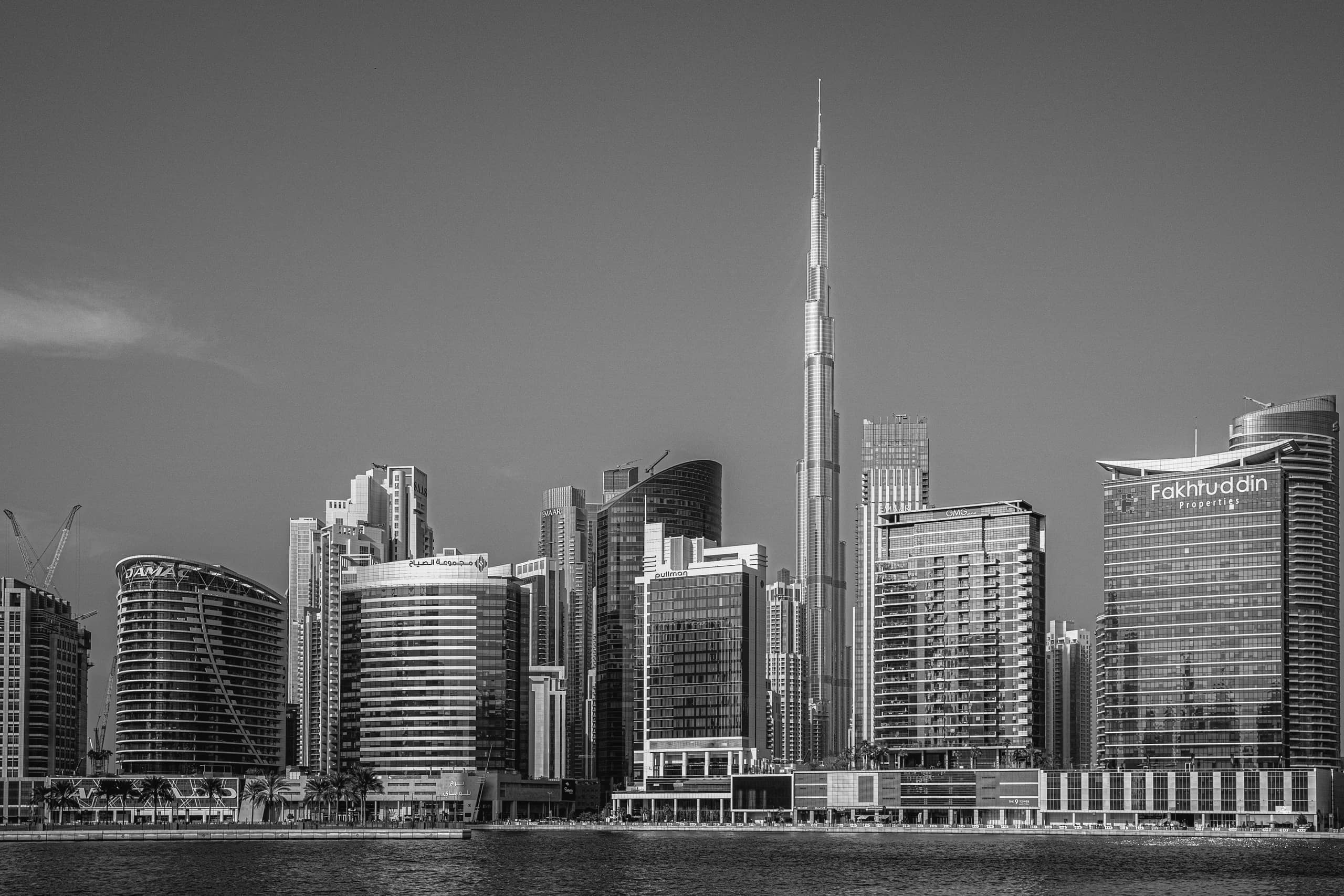 Black and white photograph of a tall skyscraper rising sharply above a crowded city skyline, viewed across a wide body of water with rippling surface texture.