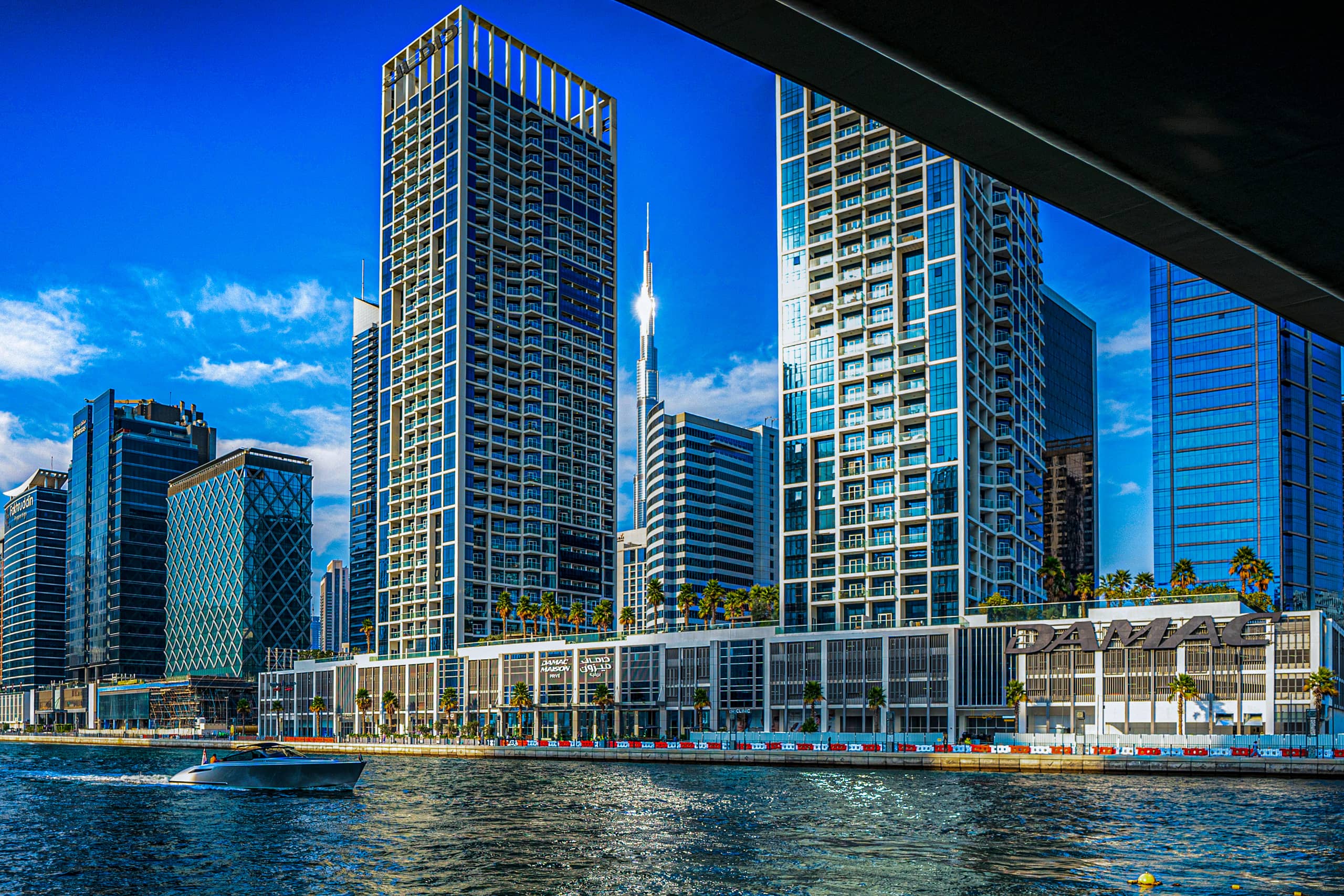 A small white motorboat creating a wake on the water in front of a modern city skyline with tall skyscrapers, viewed from beneath an overpass.
