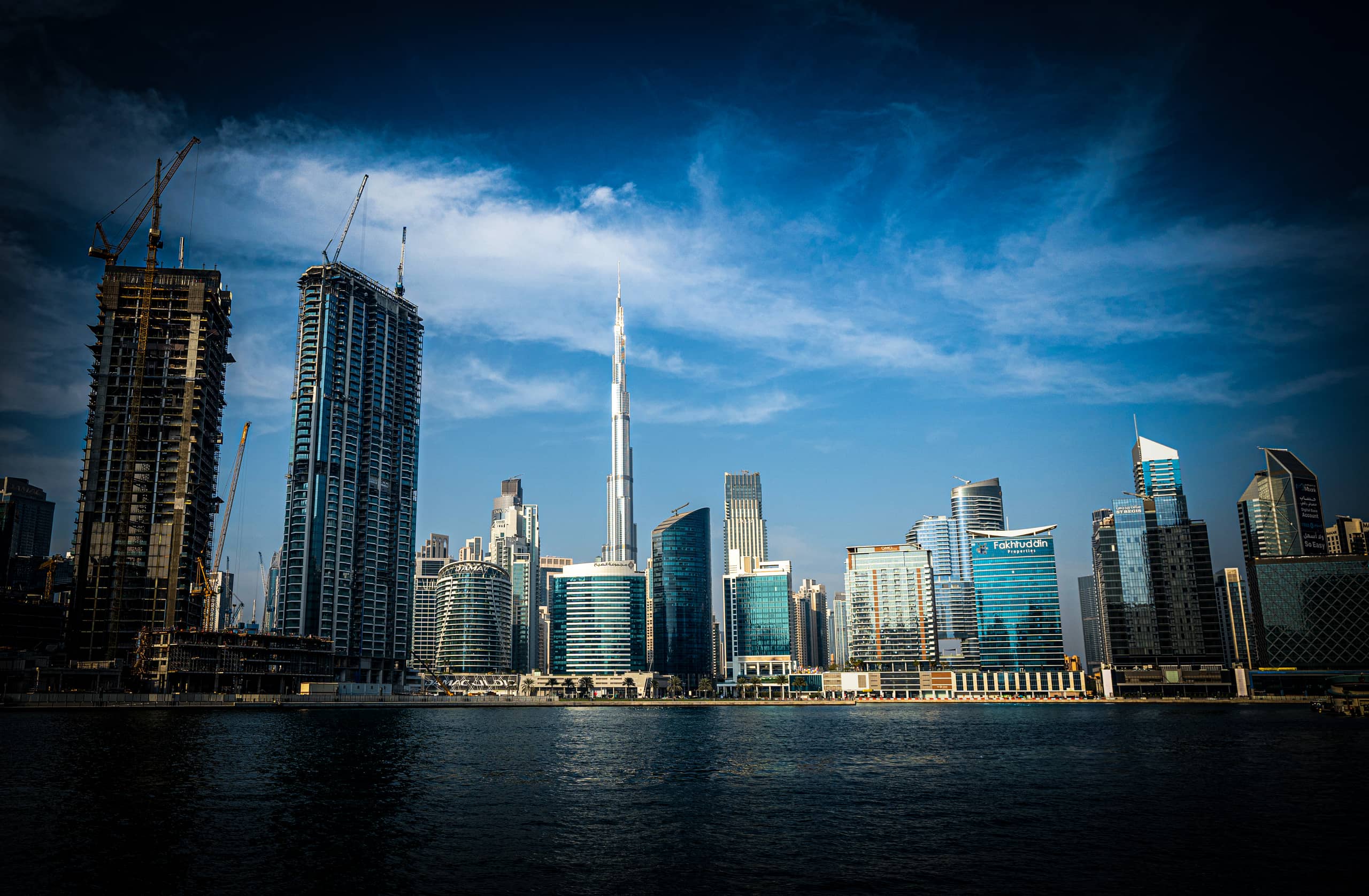Wide-angle architectural view of a metropolitan waterfront skyline featuring tall glass skyscrapers, unfinished buildings with construction cranes on the left, and a prominent central spire, all under a blue sky with soft clouds.