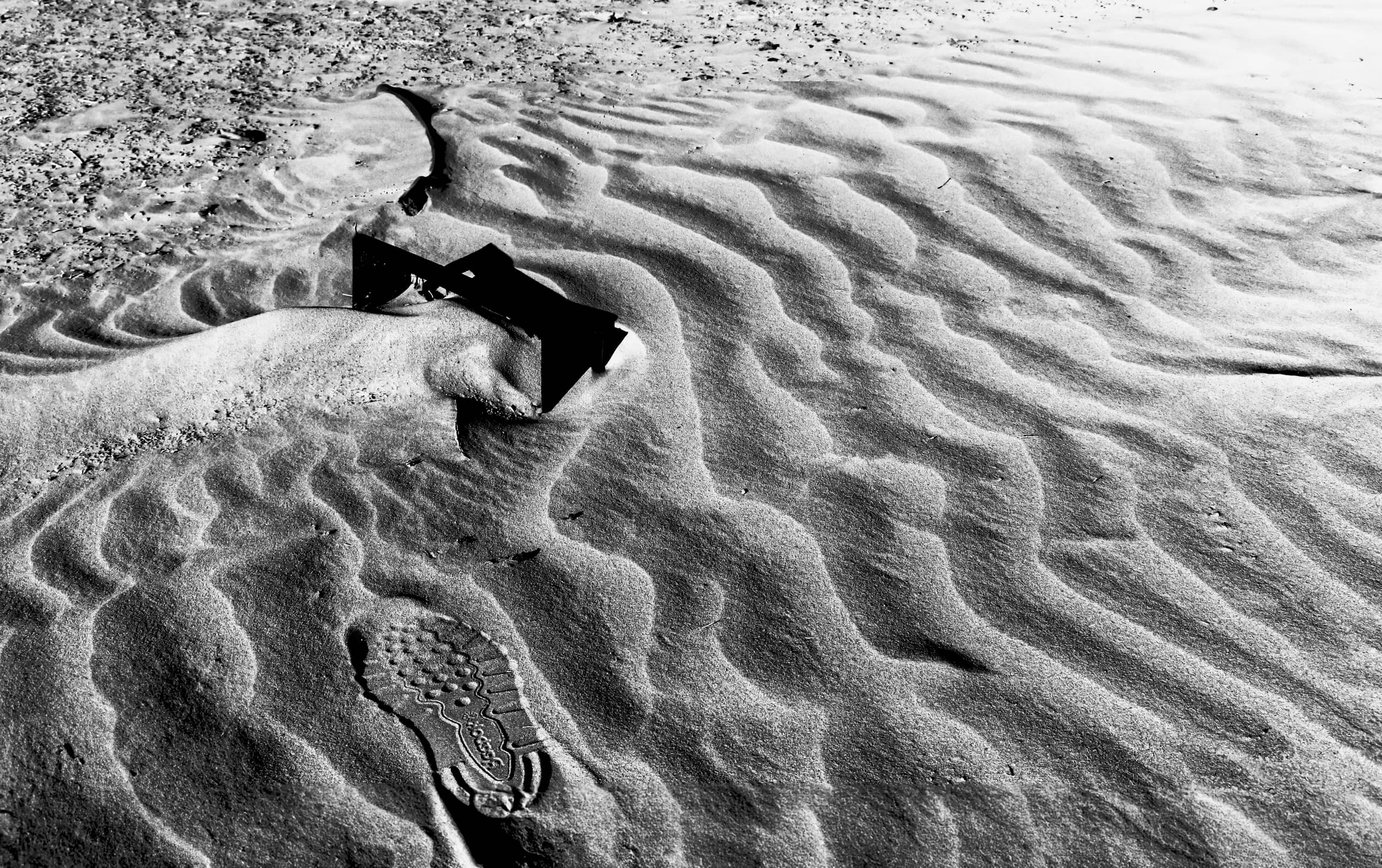 A high-contrast black and white photograph showing wind-swept sand ripples, a clear shoe tread indentation in the foreground, and a small dark angular object resting on a dune crest.