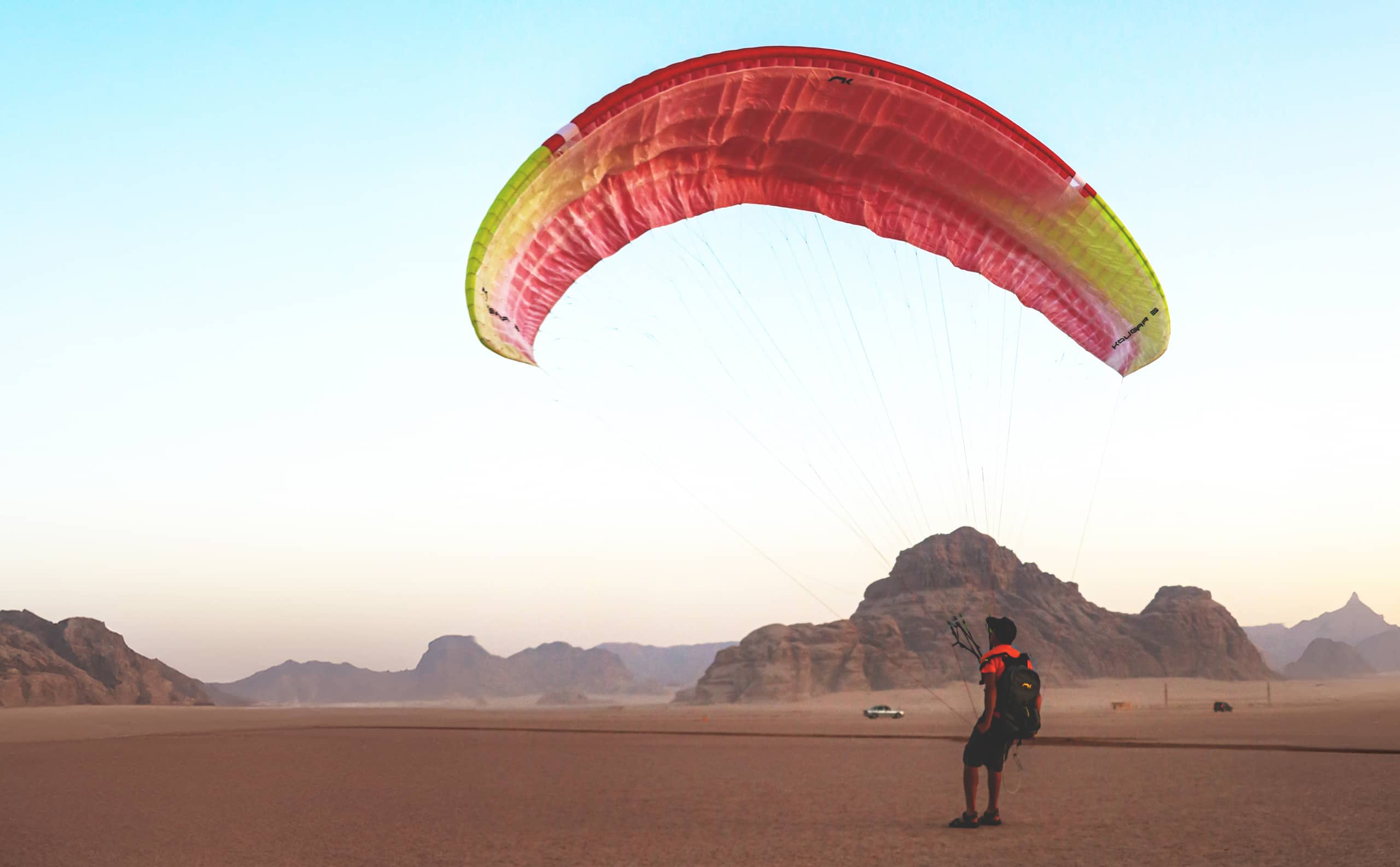 A paraglider with a large red, pink, and yellow curved canopy stands in a vast desert landscape with distant rocky mountains under a pale sky.