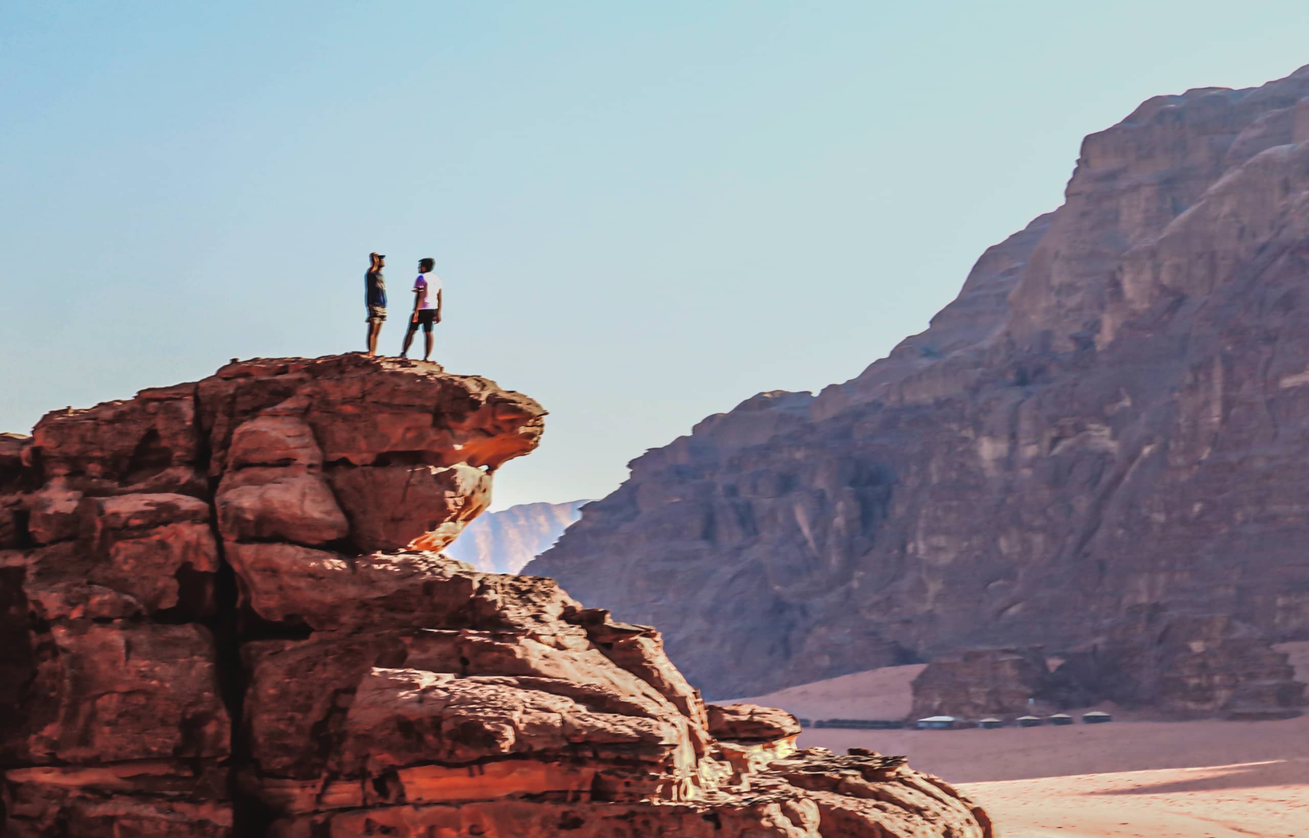 Two people standing on the rocky outcrop of a desert cliff, viewed against a vast expanse of mountains and clear sky.