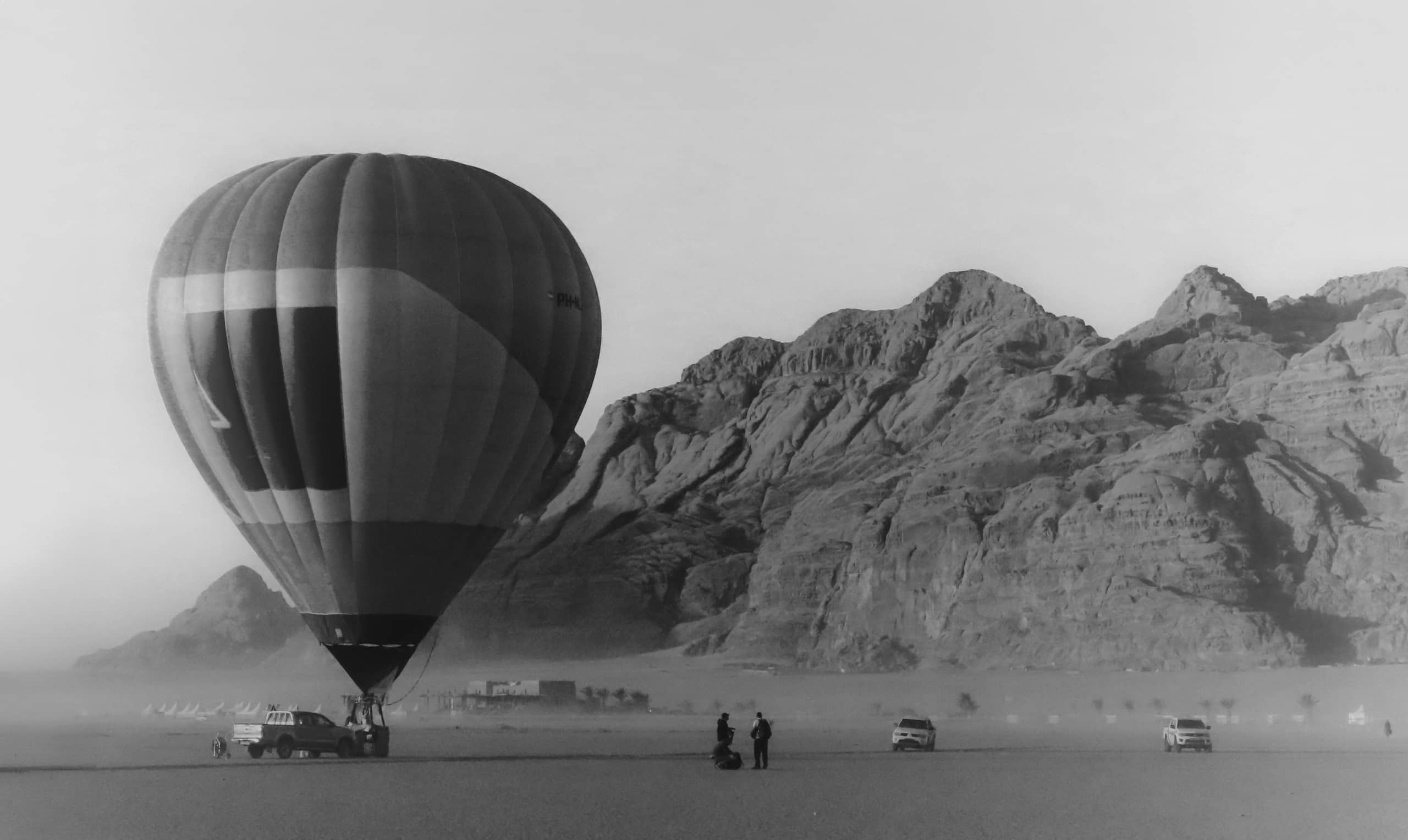 Monochrome landscape photograph featuring a large striped hot air balloon on the left, rugged desert mountains in the background, two tiny human figures standing in the lower center, and pickup trucks parked across the flat sandy ground.