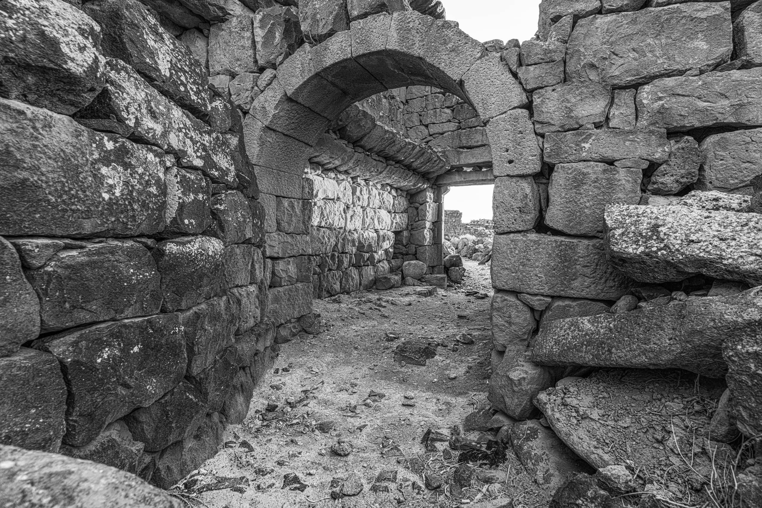 A black-and-white photograph looking down a dusty corridor formed by rugged, stacked stone walls, featuring a prominent arched doorway leading to a bright opening in the background.