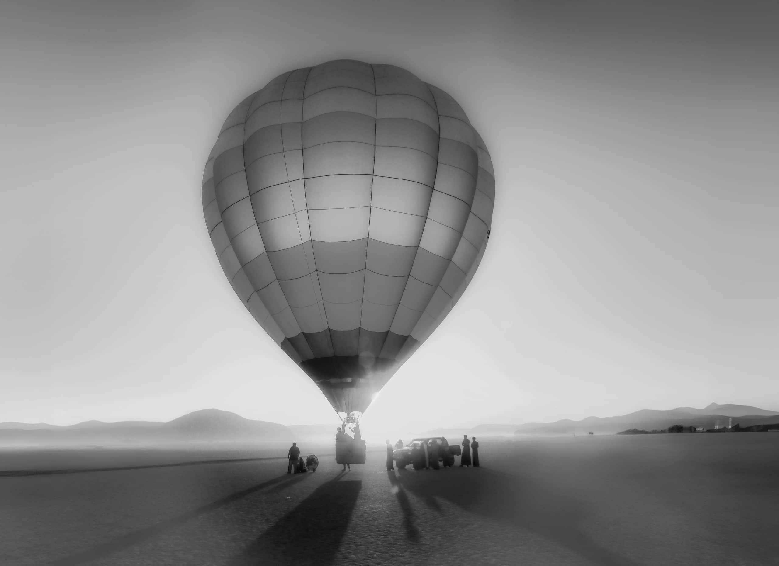 Monochrome photograph of a large hot air balloon centered in the frame with silhouetted figures and a vehicle standing beneath it on a flat landscape, casting long shadows toward the viewer.