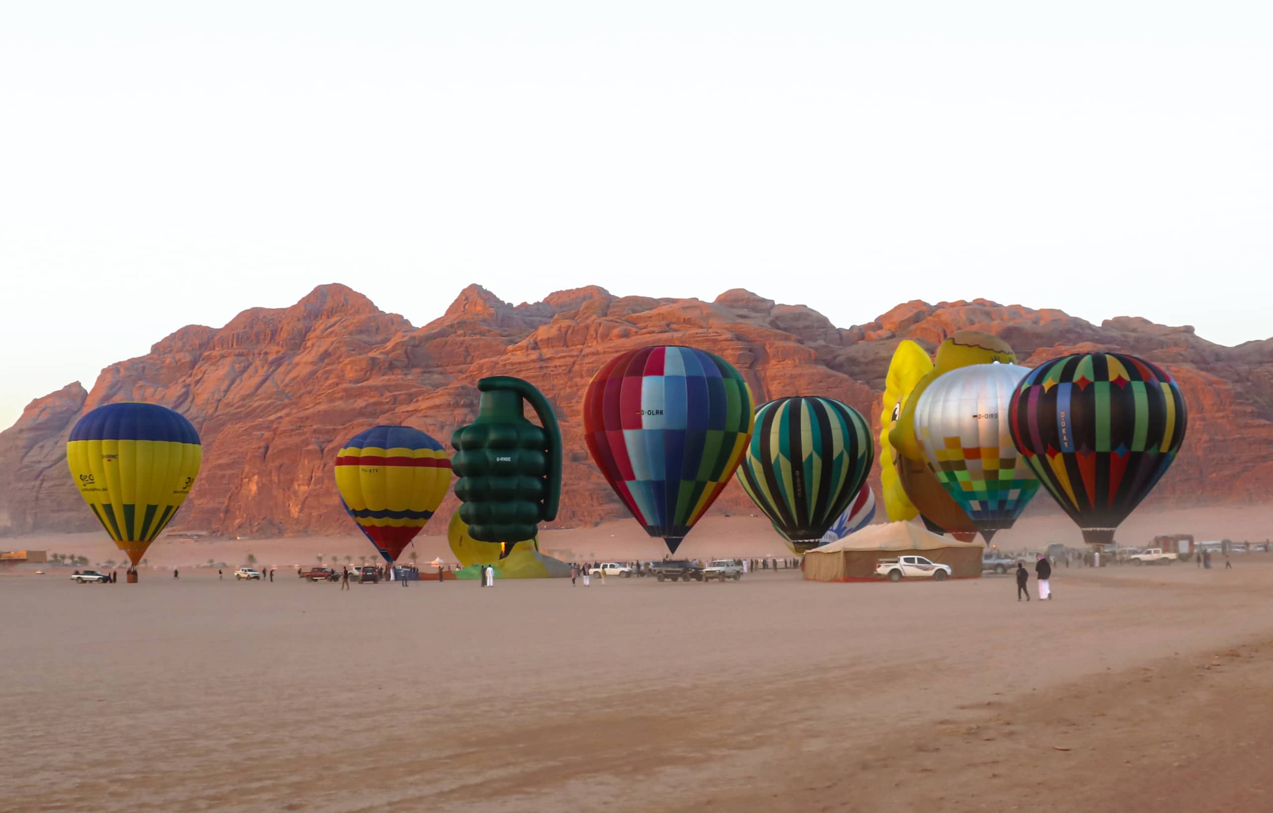 Wide-angle shot of multiple hot air balloons lined up on a flat sandy desert floor, with dramatic reddish mountain ranges rising in the background under an overcast sky.