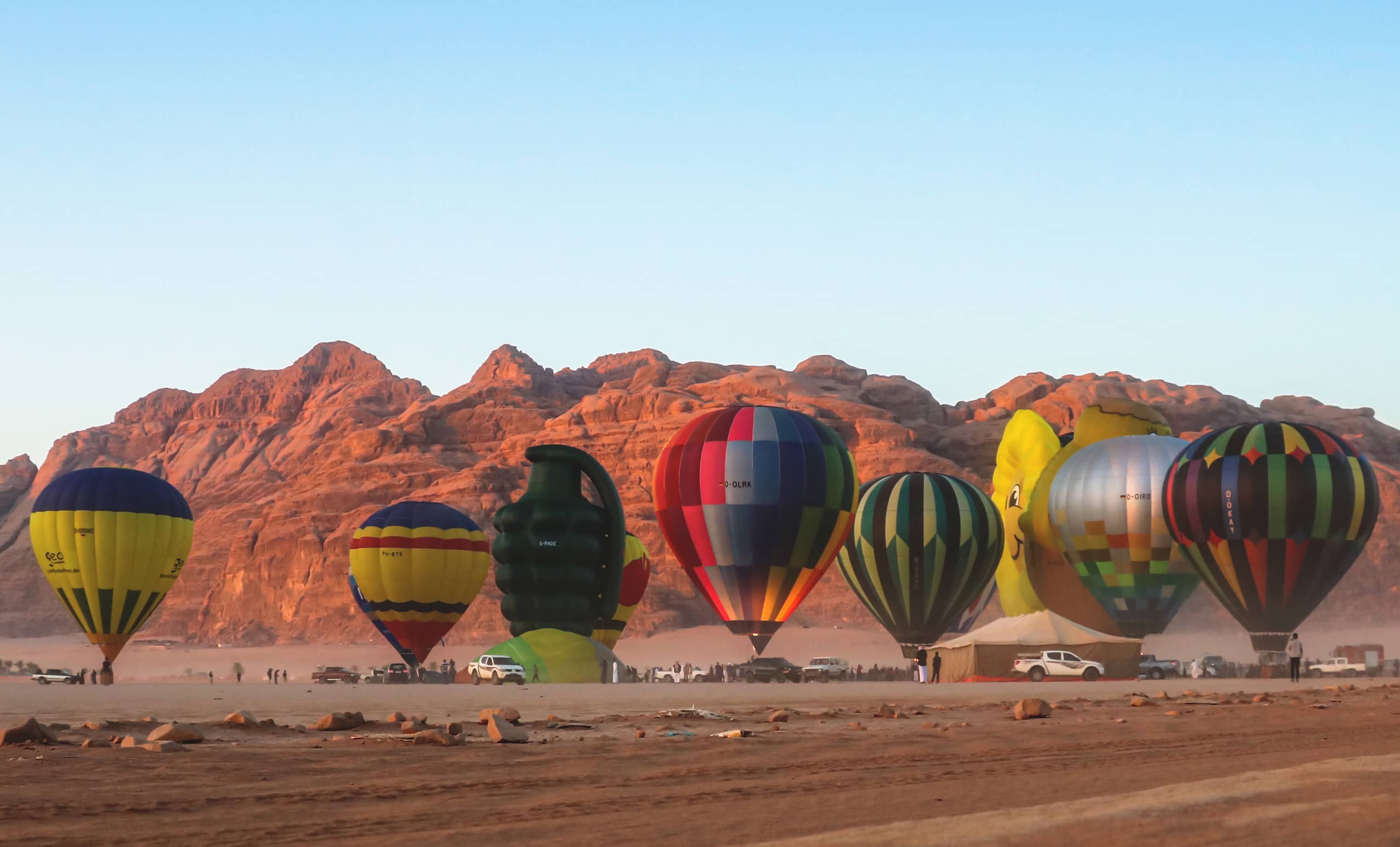 Wide shot of several large hot air balloons in various vibrant patterns lined up on the desert floor beneath a range of rugged sandstone mountains.