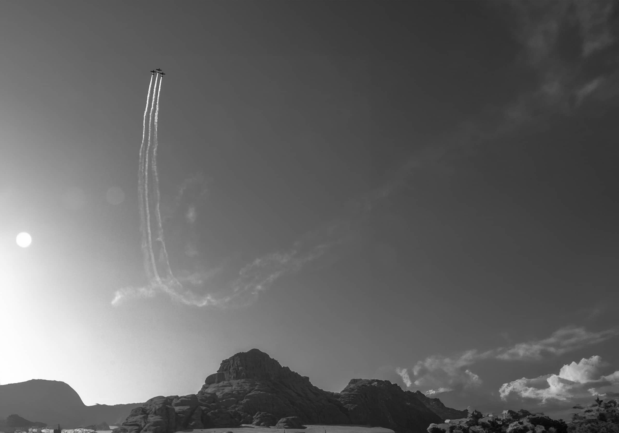 Monochrome landscape photograph featuring three small airplanes high in the sky leaving parallel white smoke trails that curve downward, with dark rock formations spanning the lower horizon and the sun shining brightly on the left.