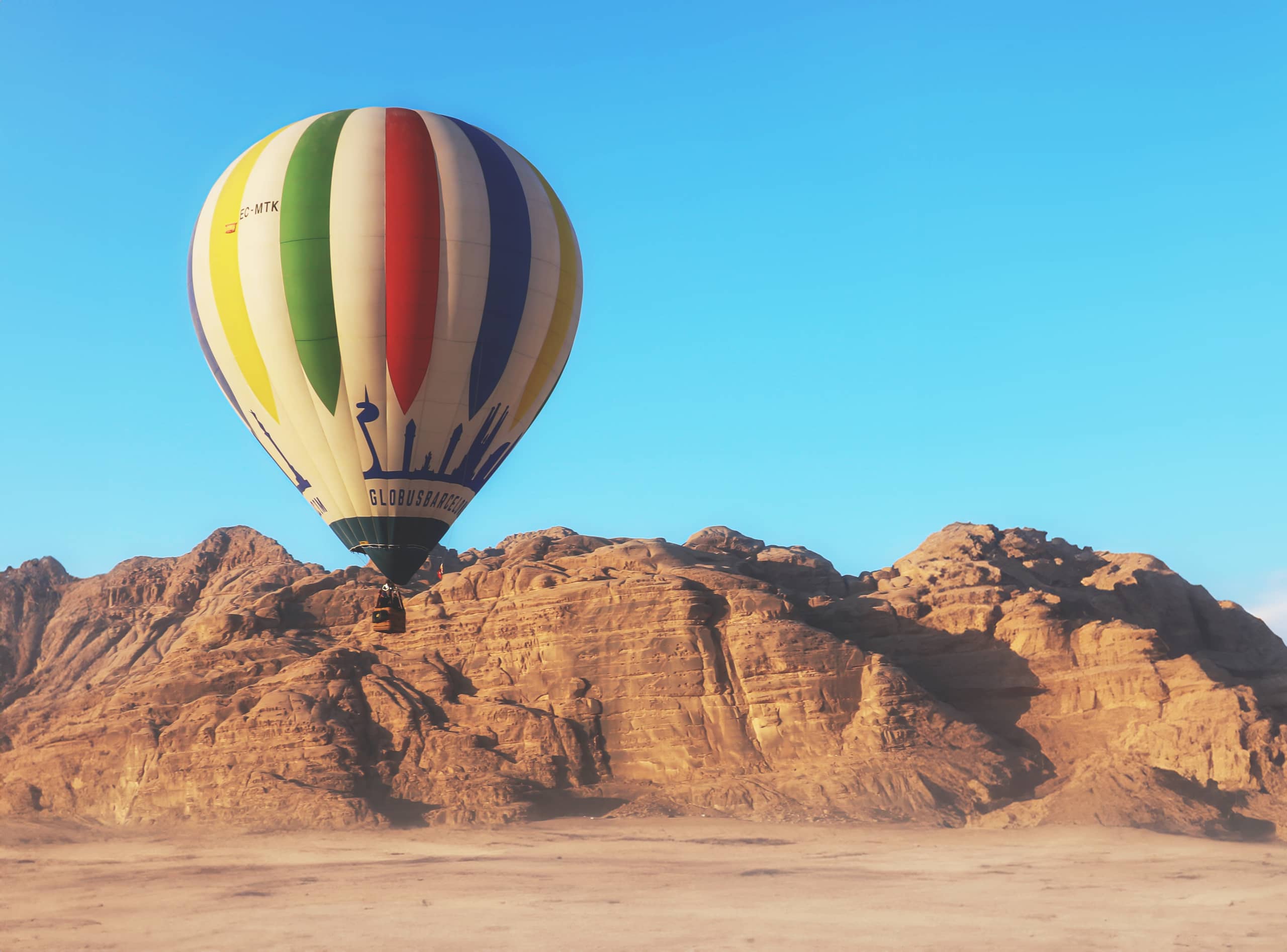 A multi-colored striped hot air balloon floats in the upper left of the frame over a low rocky mountain range and flat desert sand under a blue sky.
