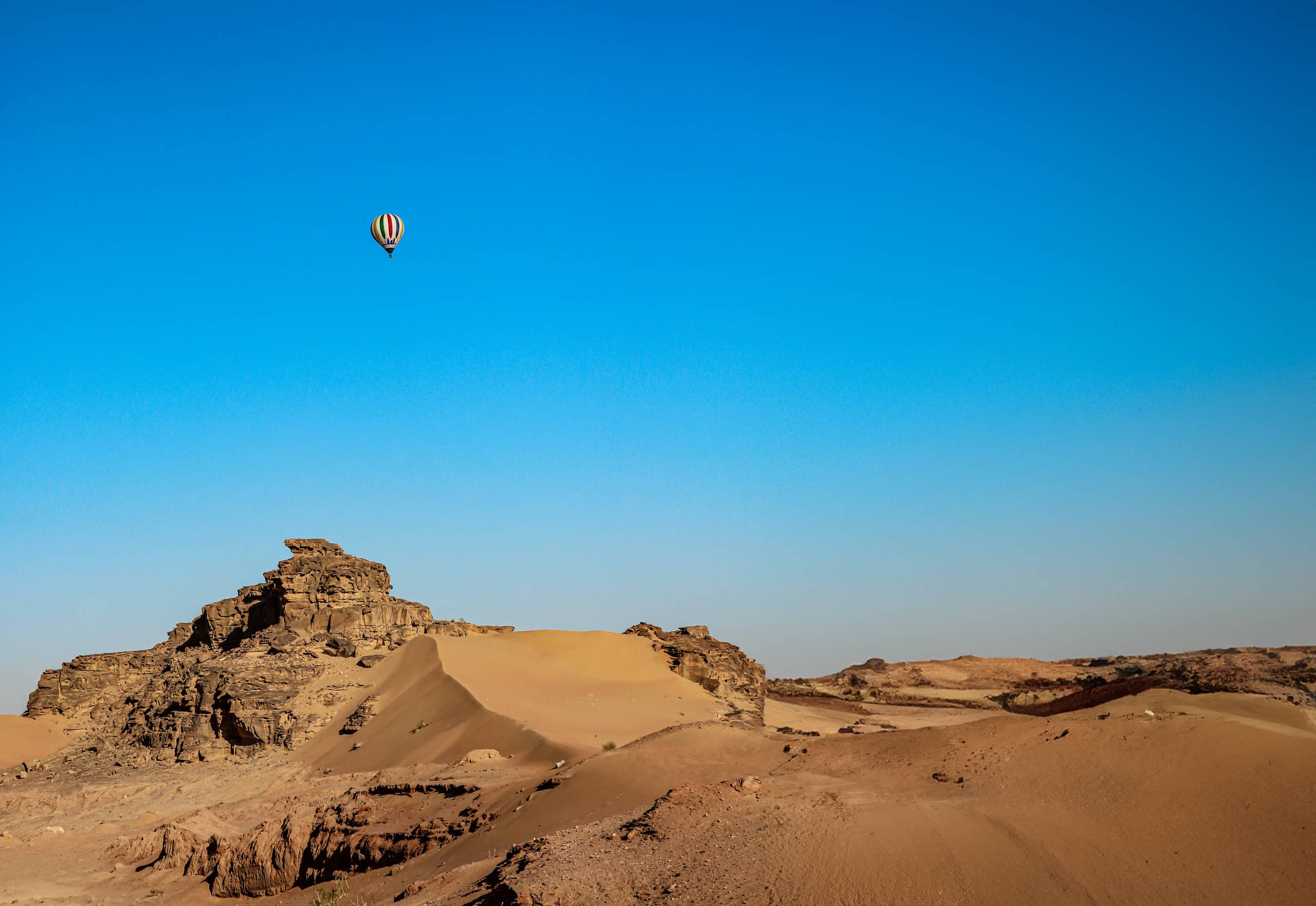 A colorful striped hot air balloon floats in a vast blue sky over a rocky desert outcrop and rolling sand dunes below.
