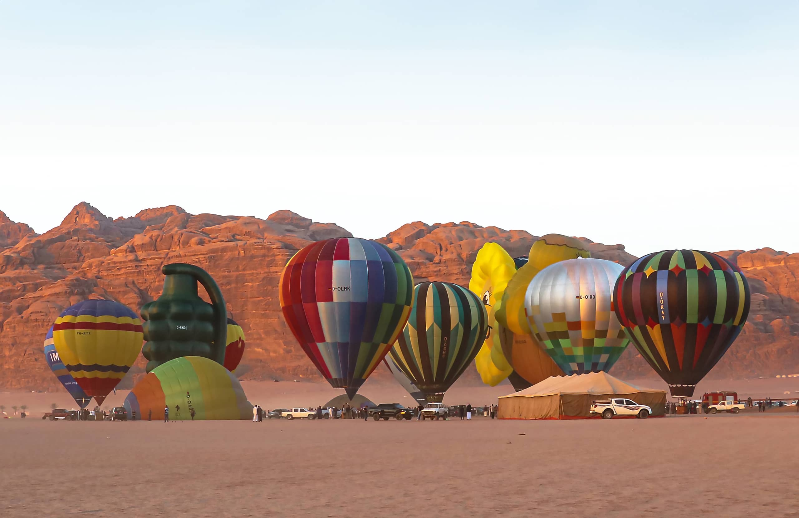 Wide shot of multiple hot air balloons in various colors and unusual shapes standing or partially inflated on a vast desert floor against a backdrop of rocky mountains under a clear sky.