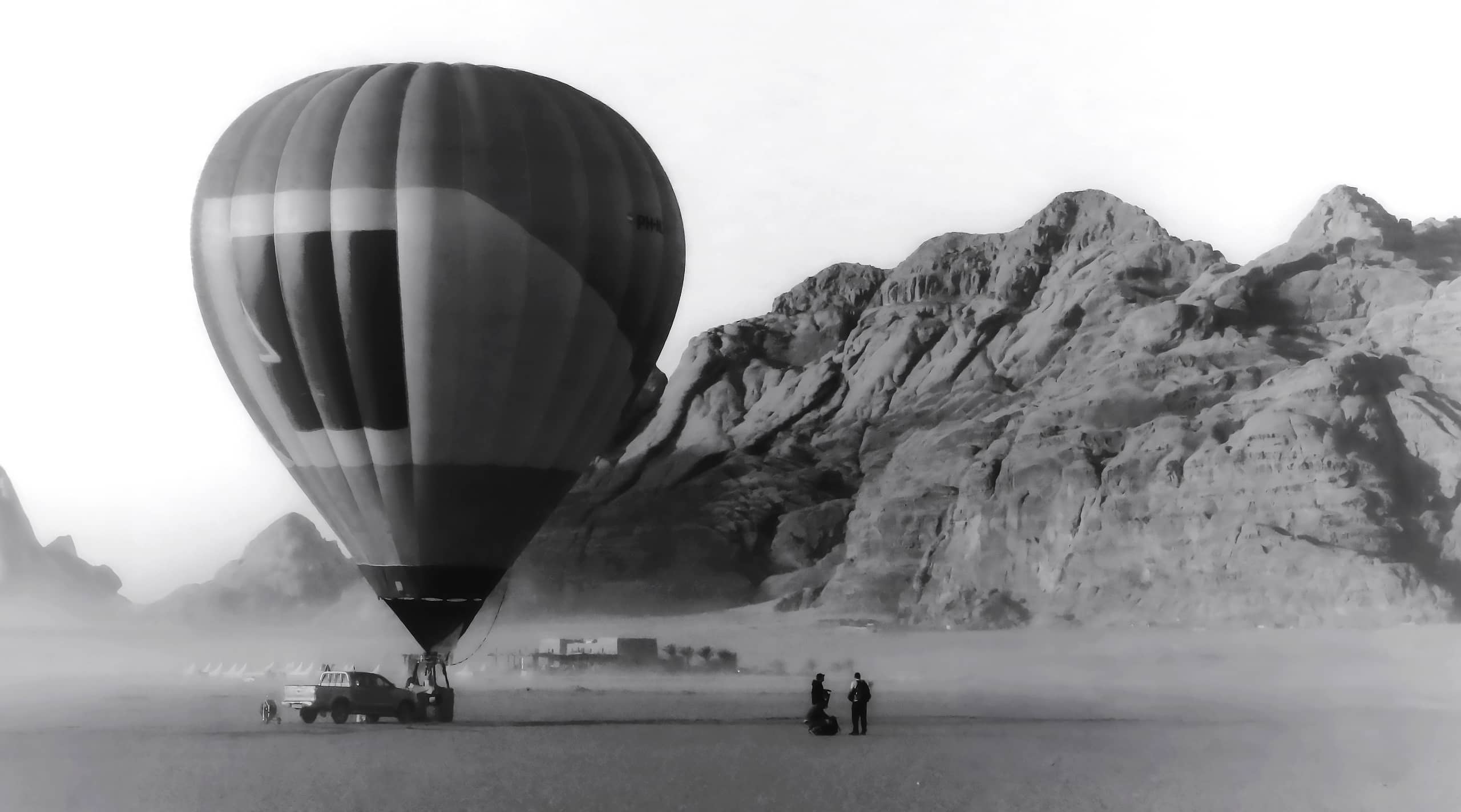 Monochrome photograph of a large hot air balloon grounded on a vast sandy plain, with two small figures standing nearby and towering rocky mountains in the background.