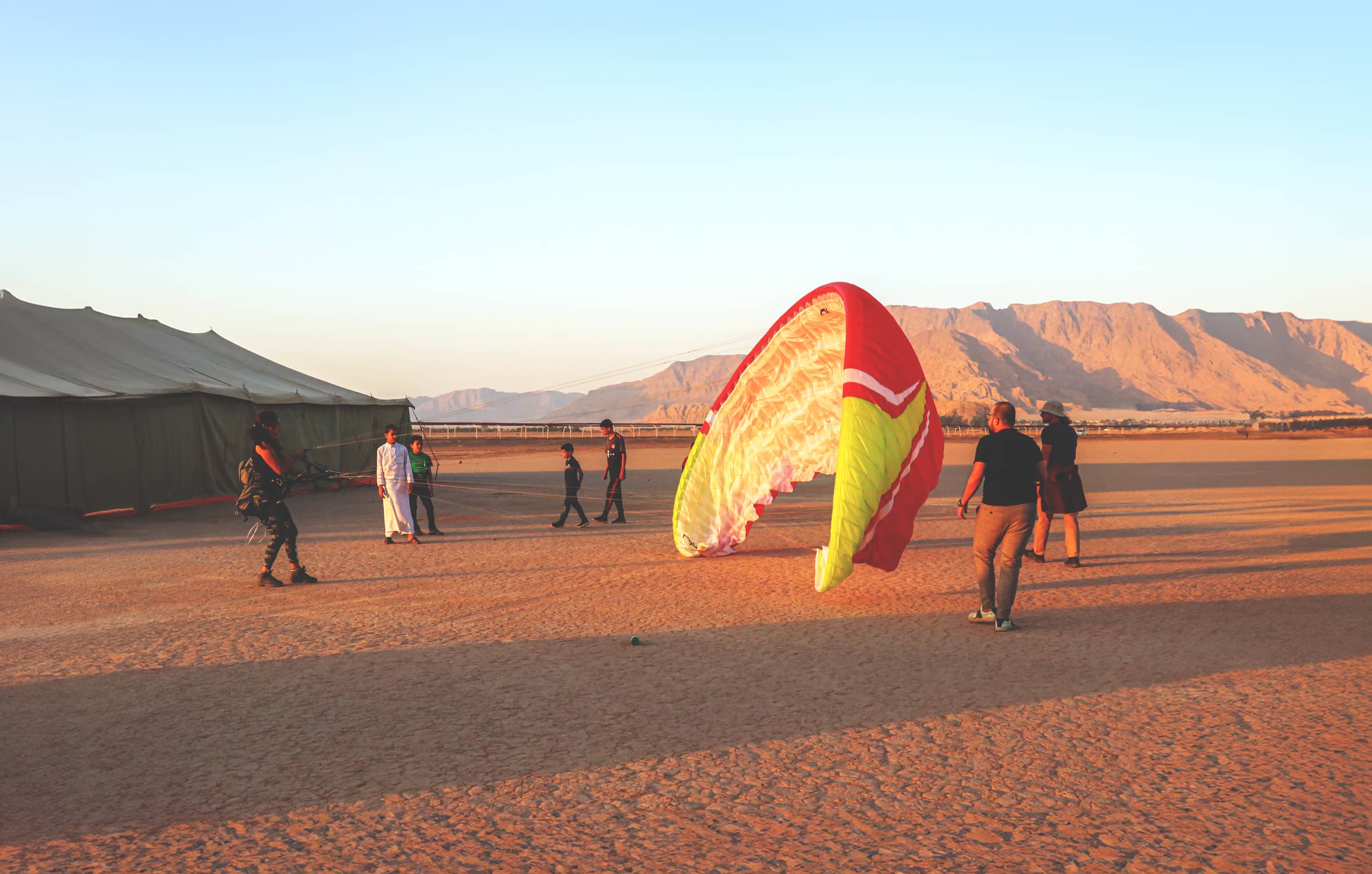 Wide-angle outdoor shot of several people standing near a large desert tent and an inflated red and yellow paragliding wing on sandy ground, with distant arid mountains under a clear sky.