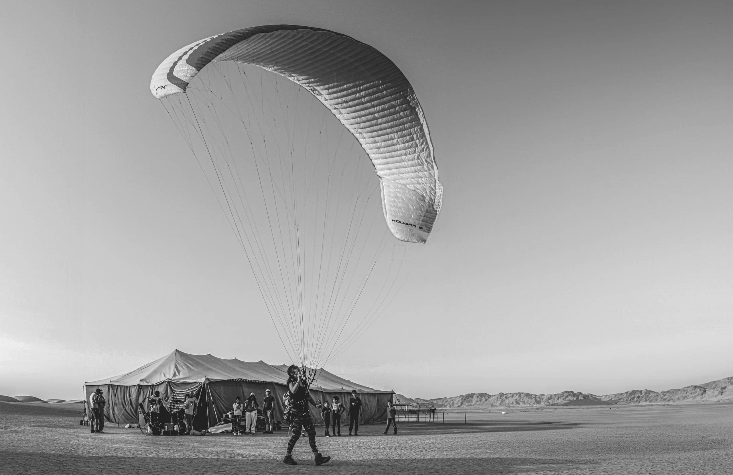Black and white photograph of a person holding the lines of a large inflated paragliding canopy in an open desert landscape, with a large tent and several distant figures visible in the background.
