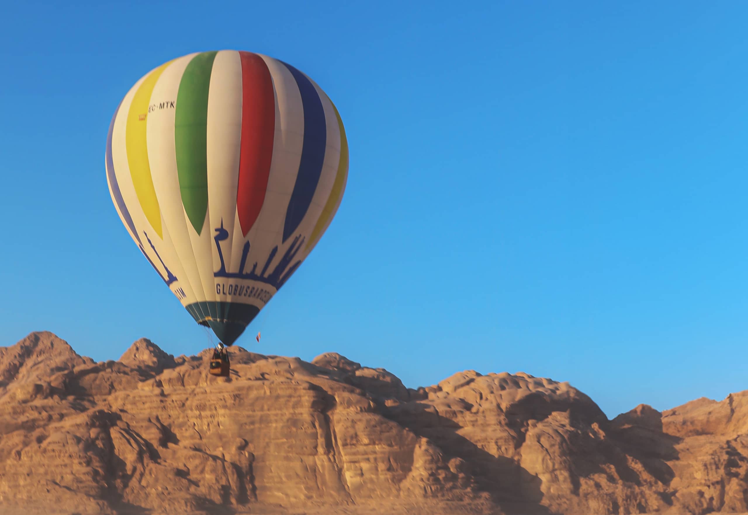 A multi-colored hot air balloon with vertical stripes of yellow, green, red, and blue floats in a clear blue sky above a rocky mountain range.