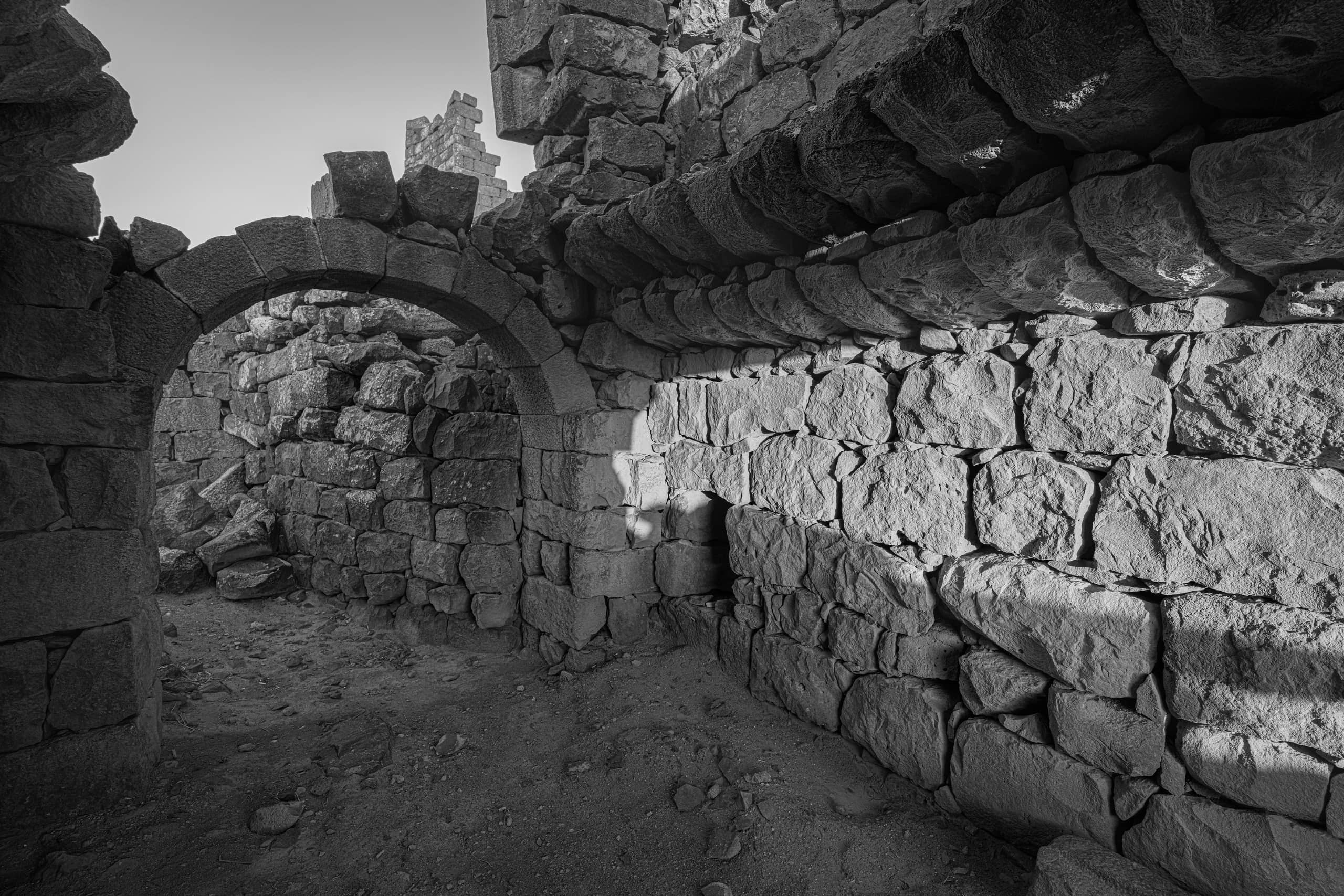Monochrome interior view of ancient stone ruins featuring a curved stone archway on the left and a large, textured masonry wall on the right illuminated by strong, low-angle light.