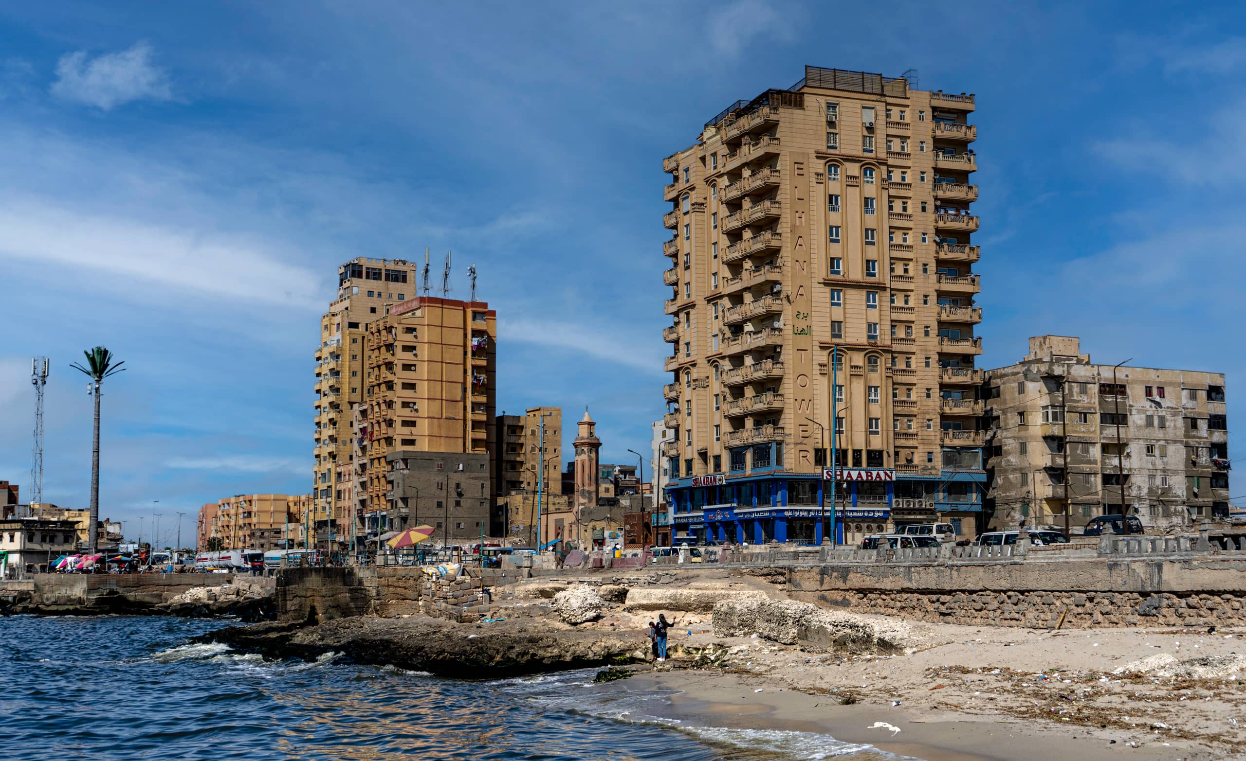 Wide-angle daytime exterior shot of tall beige apartment towers lining a rocky shore and sandy beach next to the sea, with two people standing near the water in the lower center foreground.