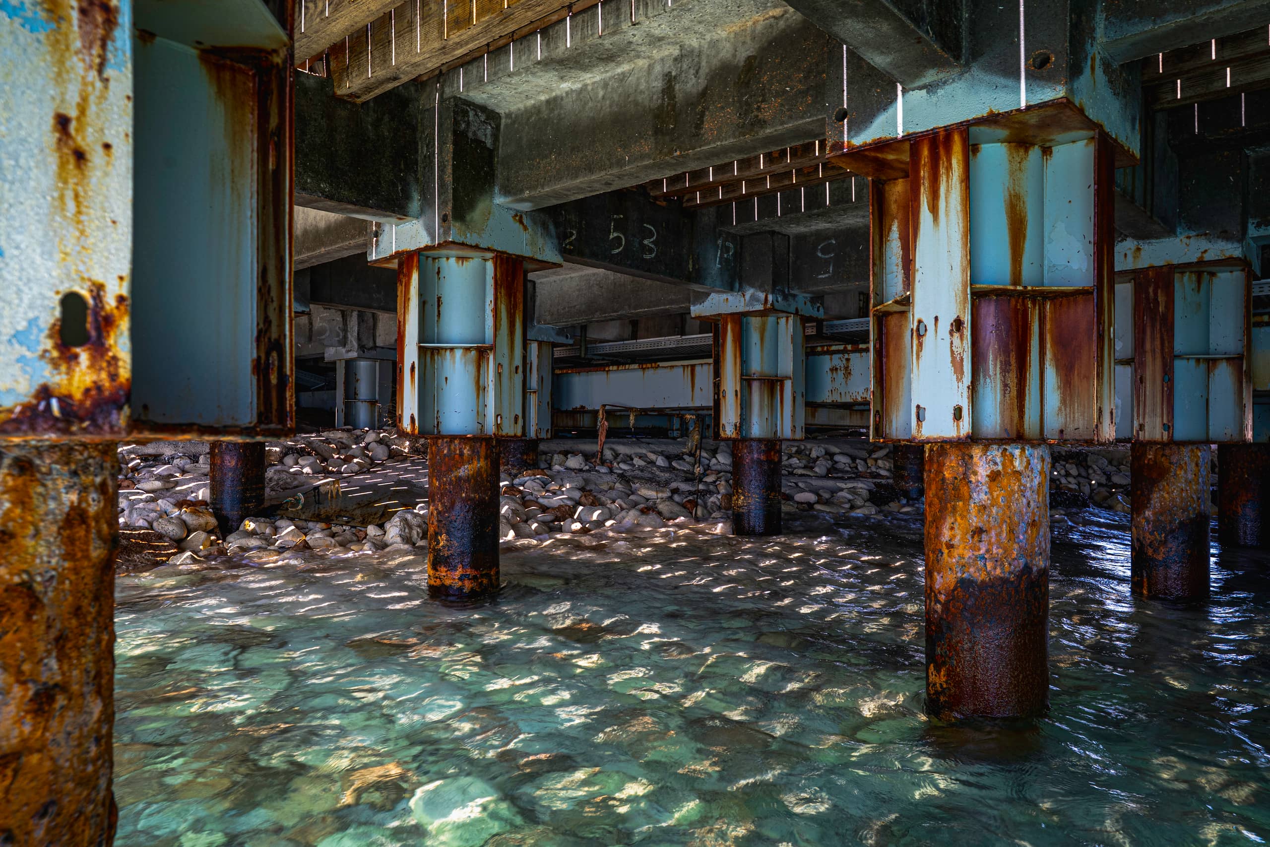 A low-angle interior view underneath a coastal pier, showing heavy rust stains on vertical steel support beams and concrete structures above shallow water rippling over smooth stones.