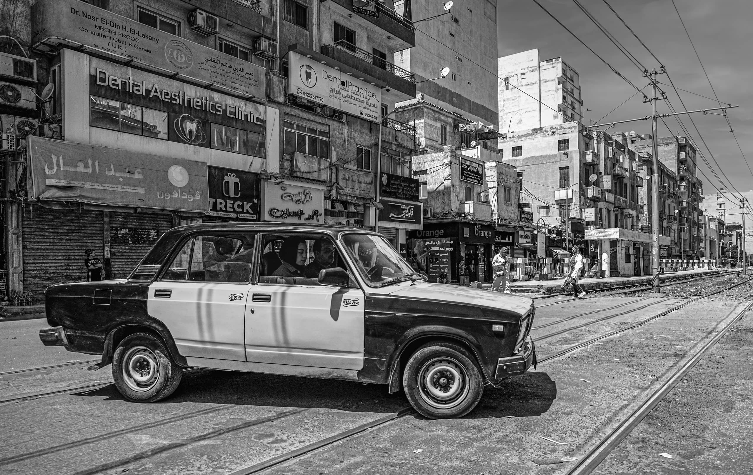 Monochrome street view of a two-tone car parked or moving alongside tram tracks, with old multi-story buildings and storefront signs lining the street in the background.