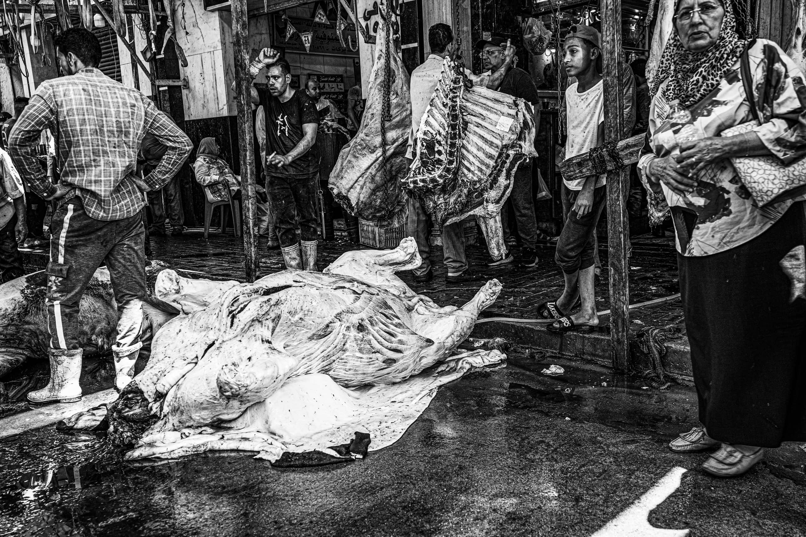 Monochrome street-level view of a bustling market stall with hanging carcasses and a large skinned hide resting on a wet concrete floor, surrounded by several figures standing and observing.
