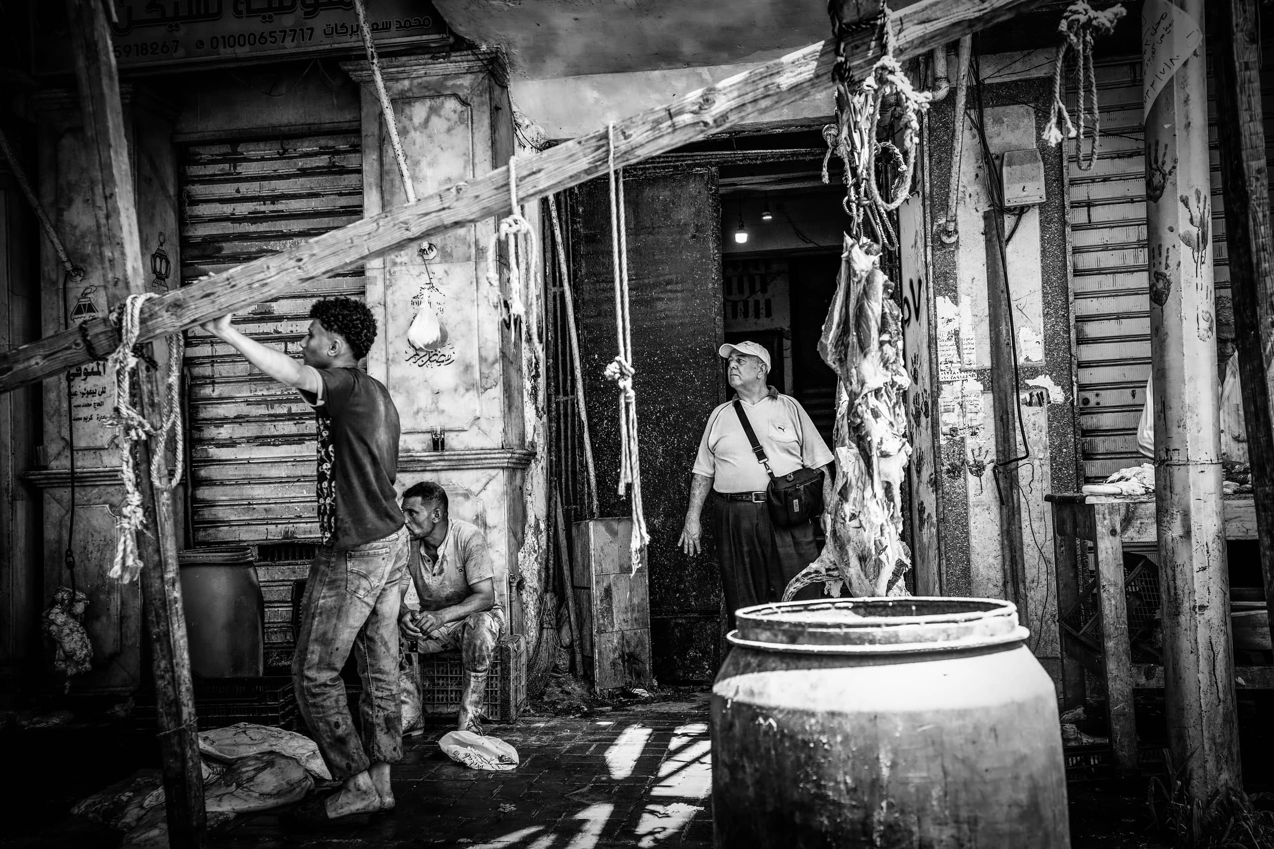 Black and white street photograph of an outdoor market scene in Alexandria, showing a young worker holding a wooden beam on the left, a seated man behind him, and an older man standing in a doorway near hanging ropes and a large plastic drum.
