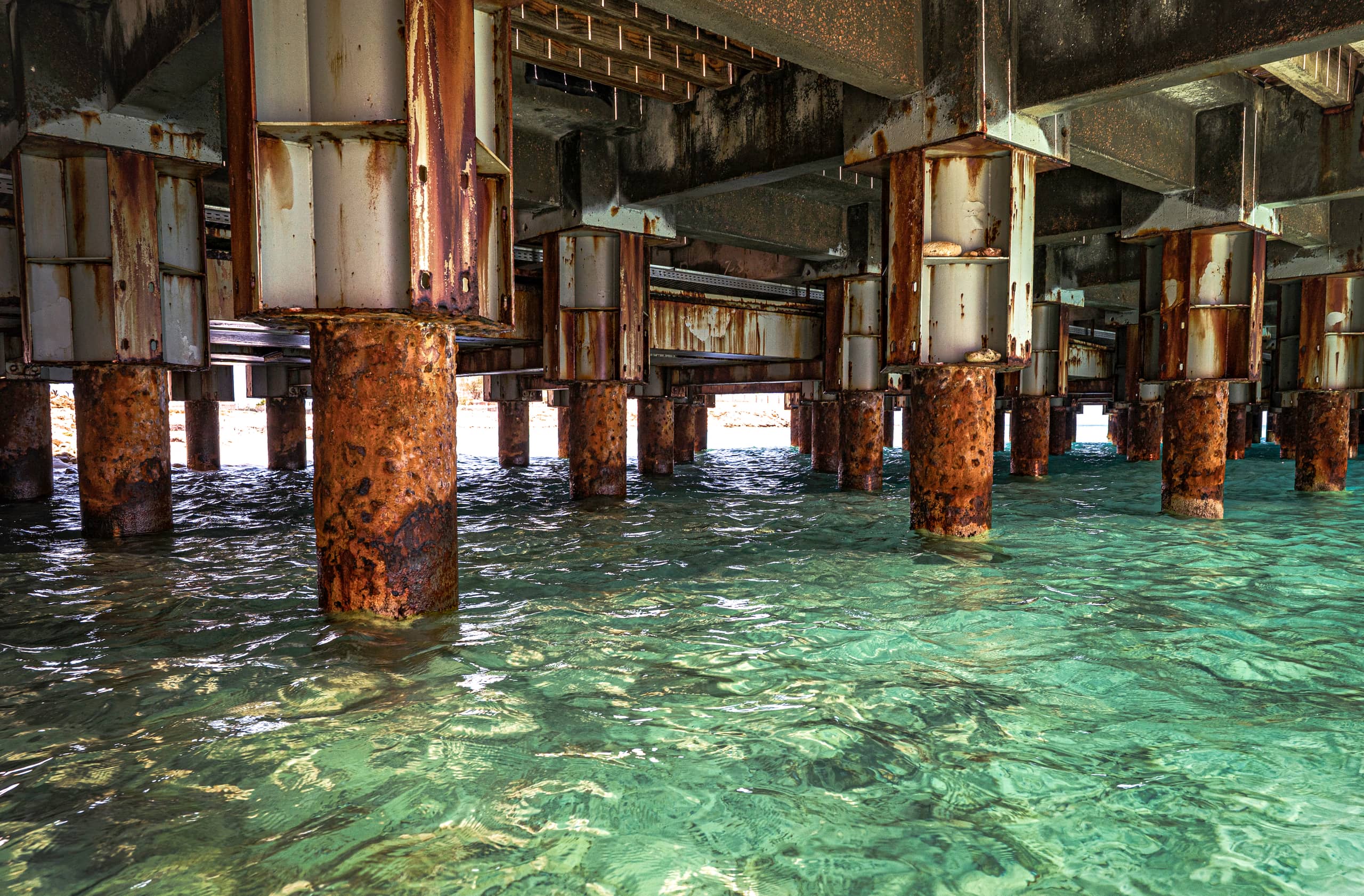 A low-angle shot underneath a concrete and steel pier, showing weathered rust-stained vertical pilings rising out of vibrant turquoise and green seawater.