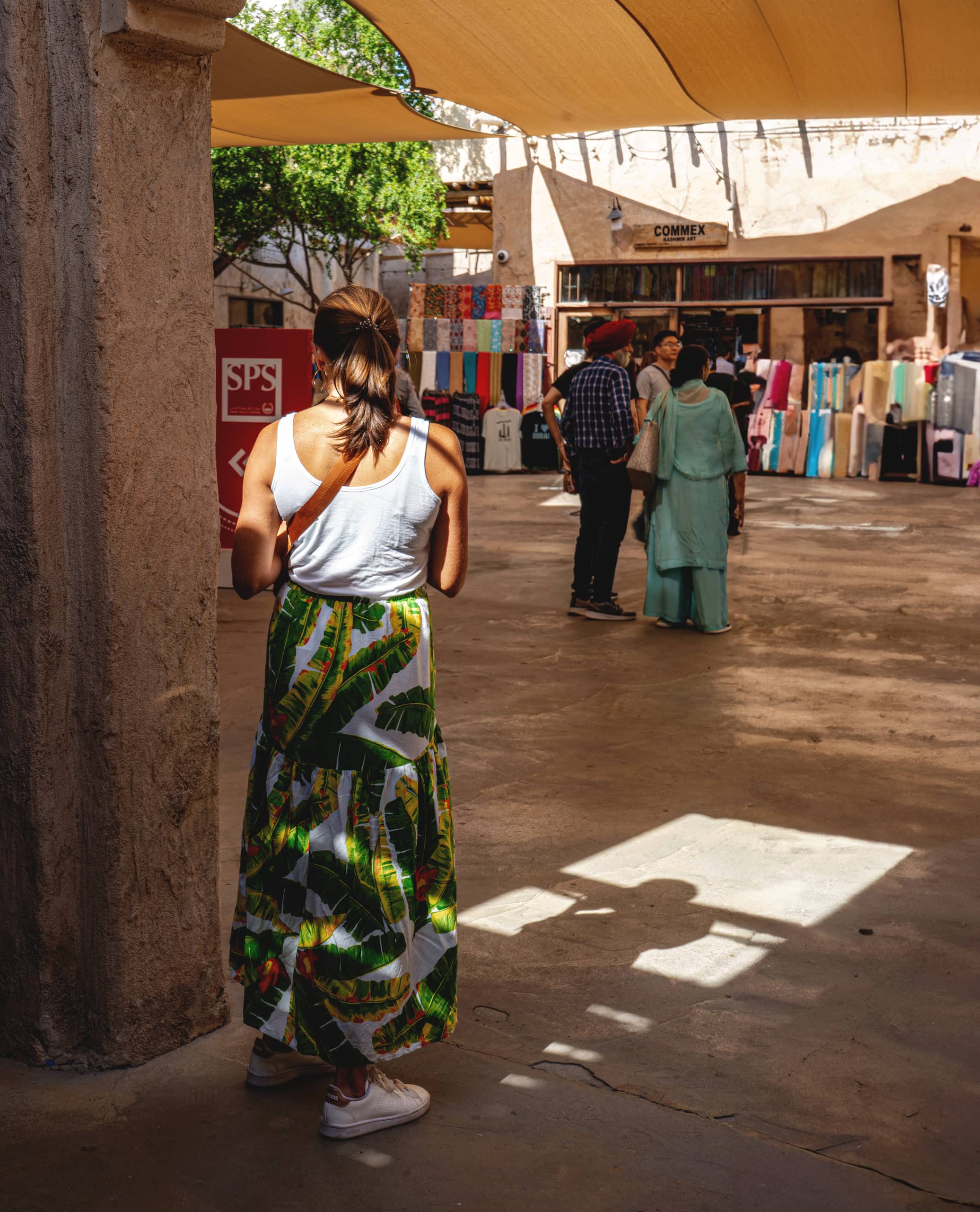 A woman viewed from behind wearing a white tank top and a patterned green and white skirt, standing in a shaded courtyard with people and shop displays visible in the sunlit background.