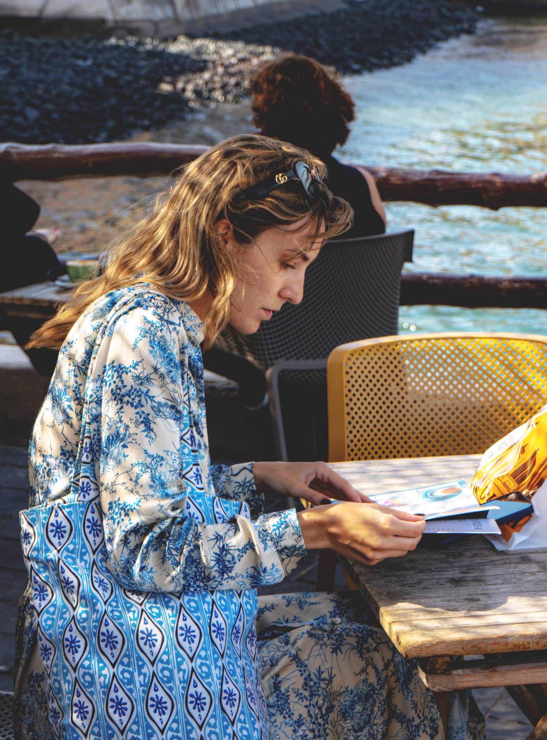 A woman with blonde hair seen in profile at an outdoor café table by the sea, looking down at a menu held in her hands while wearing a blue-and-white patterned outfit.