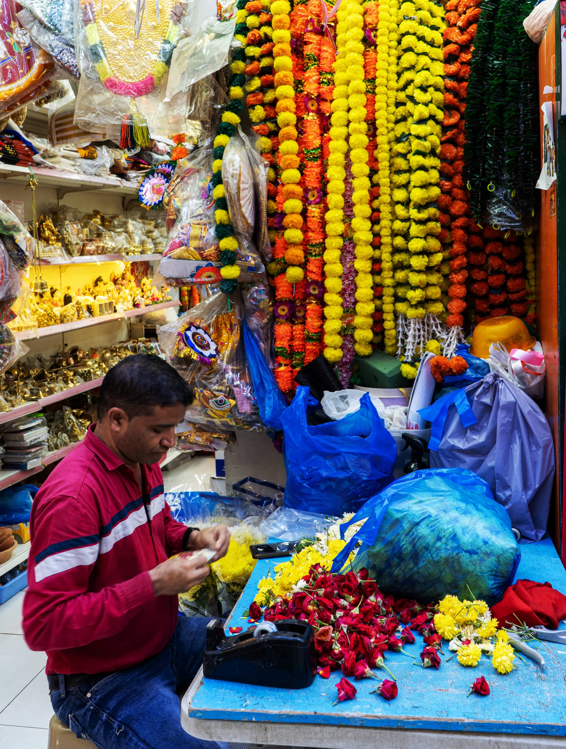 A man wearing a red collared shirt sits indoors at a bright blue table sorting flowers, surrounded by hanging floral garlands and shelves stocked with merchandise in Dubai.