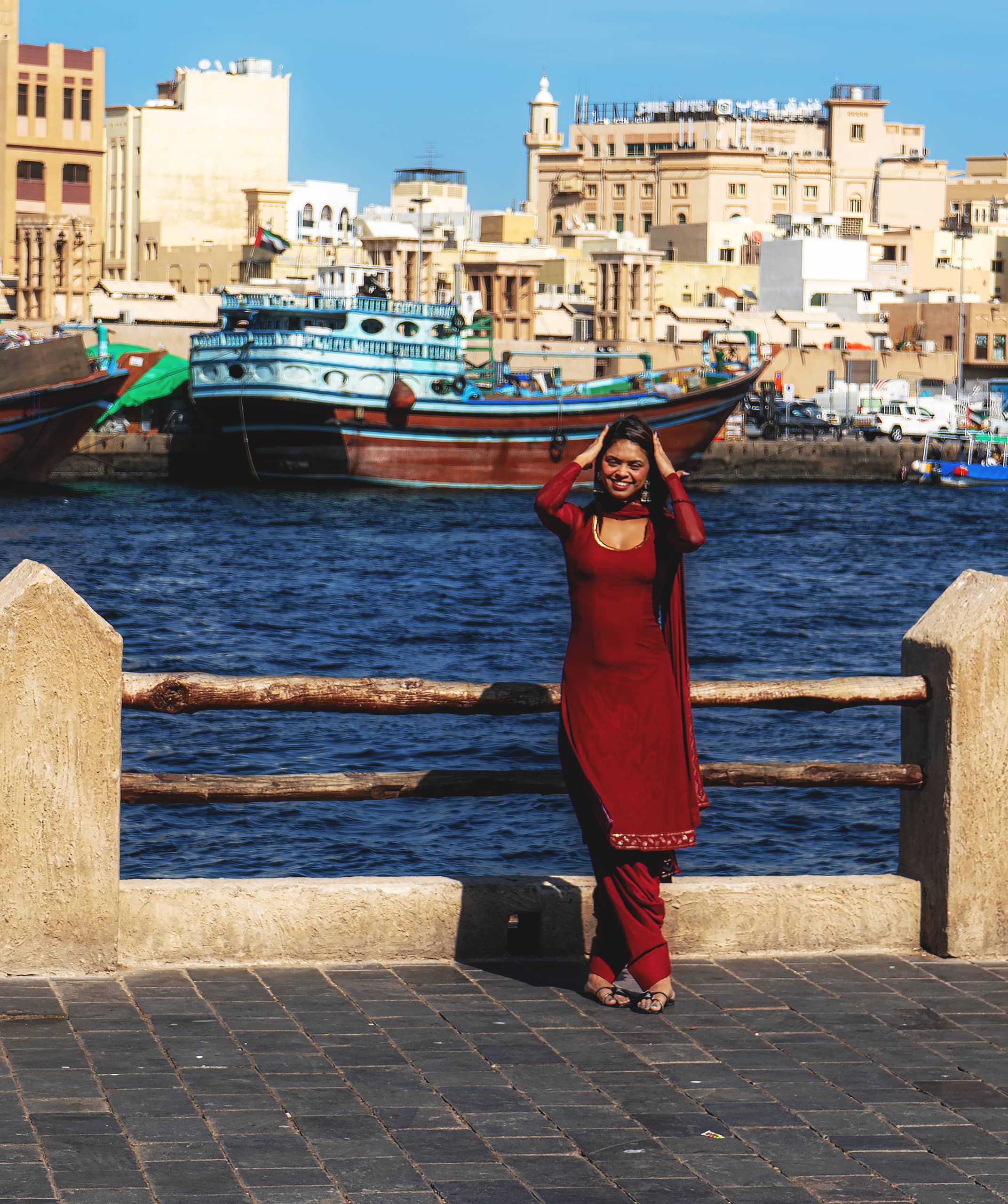 A smiling woman wearing a deep red salwar kameez poses against a low stone and log railing on a paved promenade, with a blue waterway and traditional wooden boats and historic buildings in the background under clear daylight.