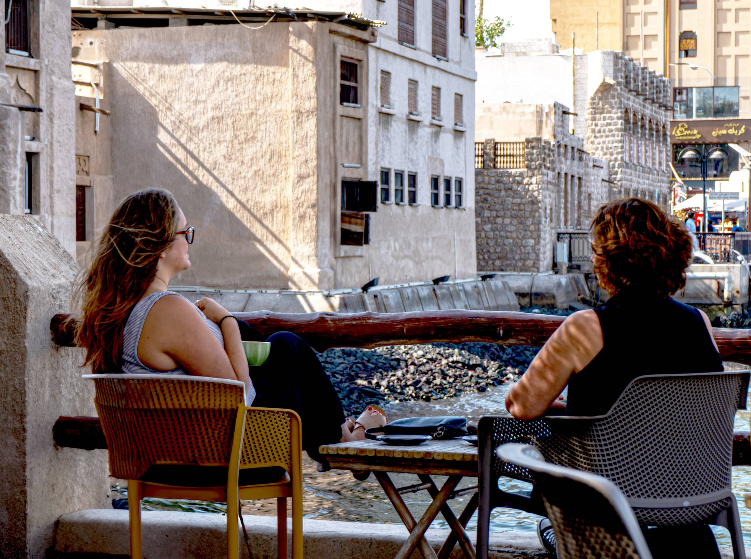 Two women seated outdoors in chairs facing each other across a small table near a stone wall and water, viewed from the side and back.