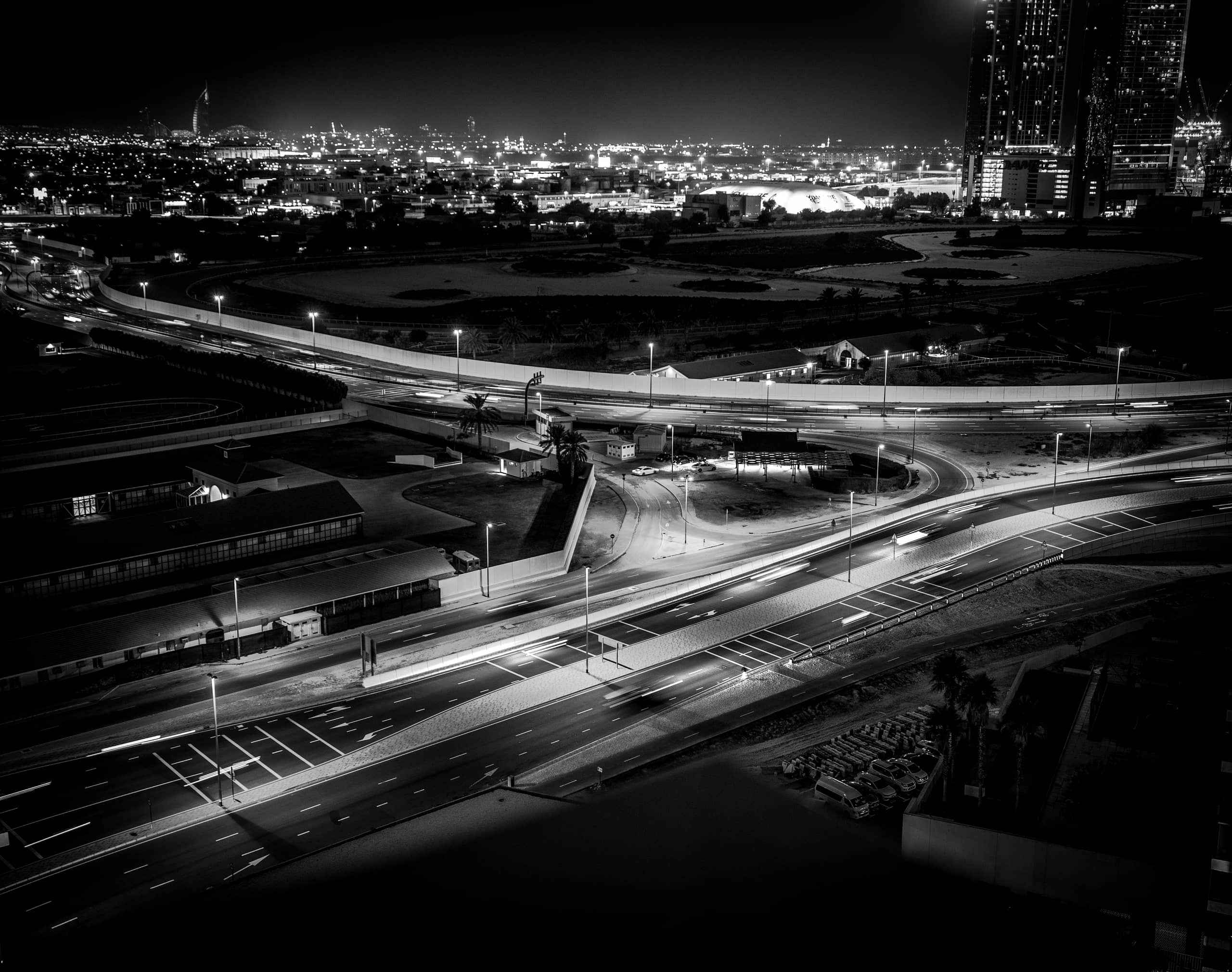 High-angle black and white night shot of urban roads with long-exposure light trails from cars, buildings, and distant city lights.