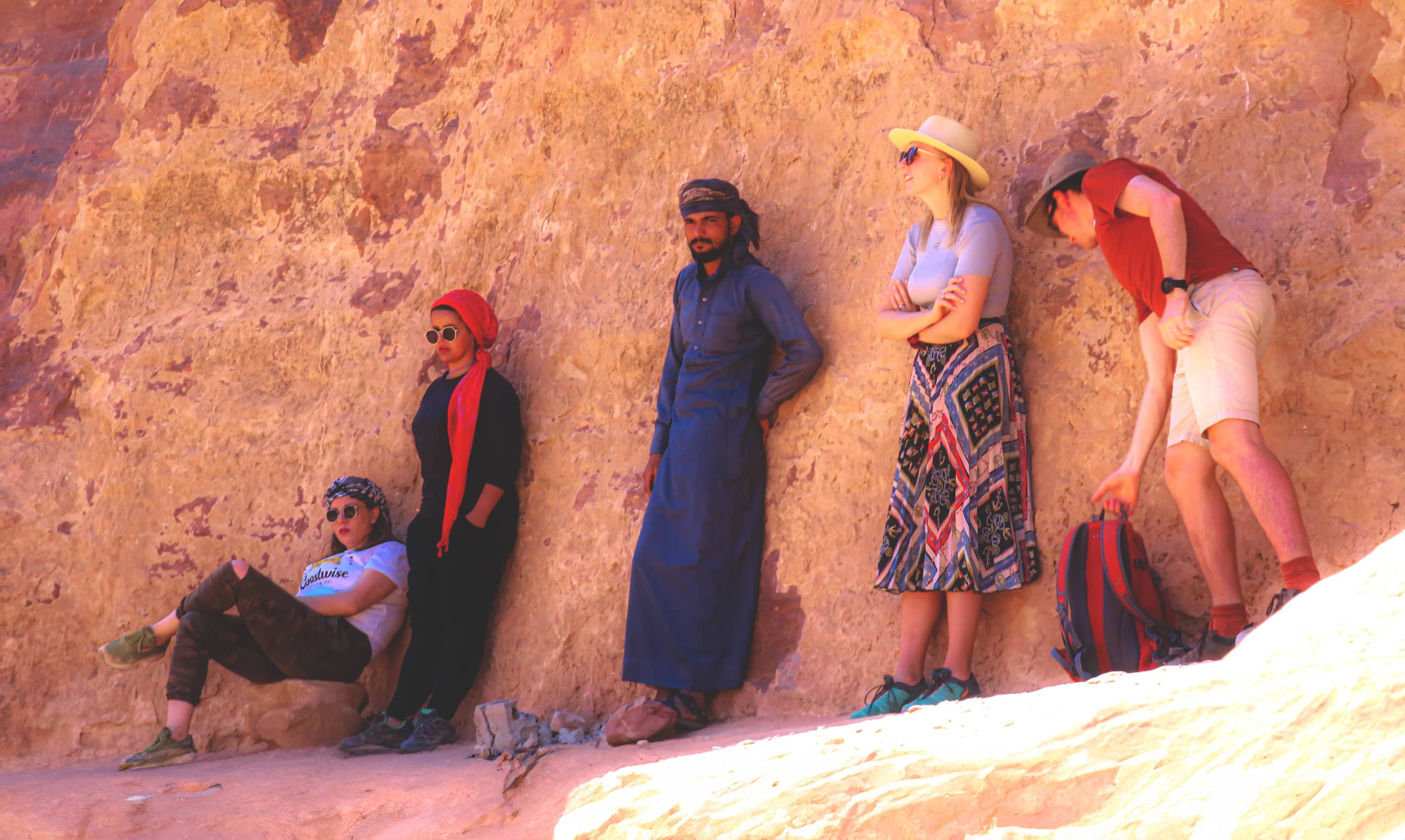 Five people leaning and sitting against a massive, weathered sandstone cliff face, varying in clothing style from casual shirts and skirts to traditional garments and headwraps.