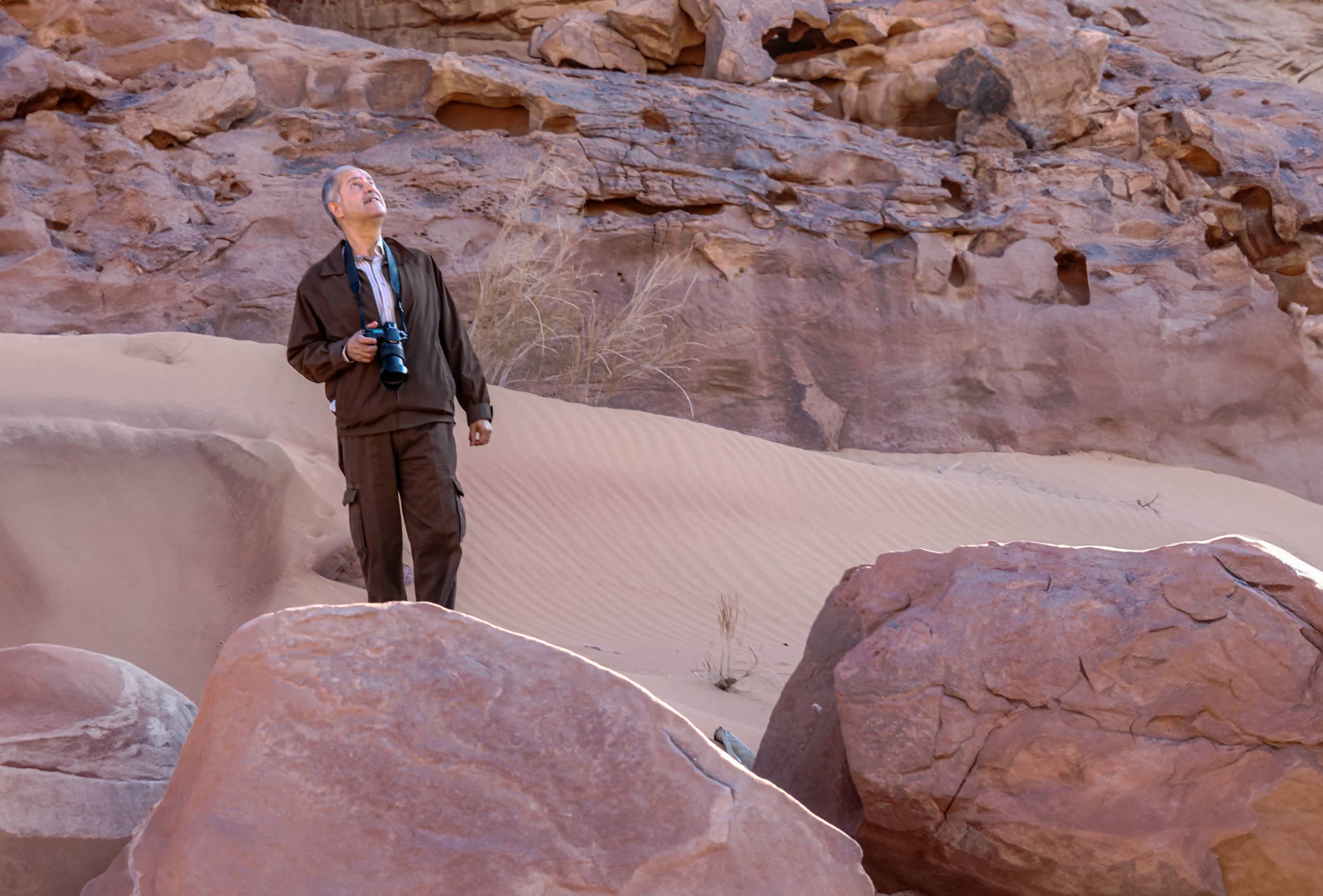 A middle-aged man with a camera slung around his neck stands on a sandy incline among large red sandstone rocks, looking up and away from the camera.