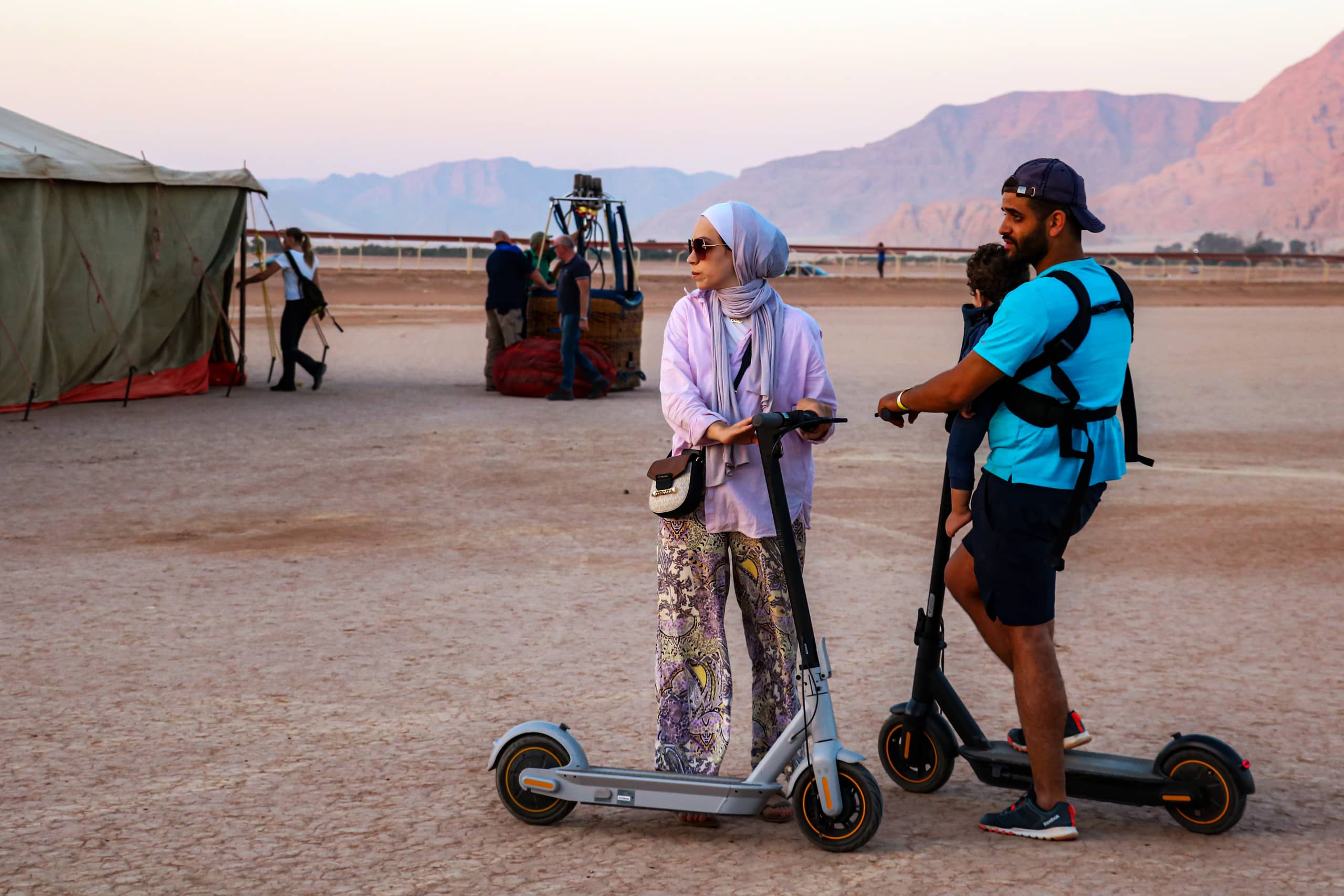 A woman wearing a hijab and patterned trousers and a man carrying a young child in a harness both stand on electric kick scooters on a dusty, dry plain outdoors, with a tent, hot air balloon basket, and distant mountains in the background under soft daylight.