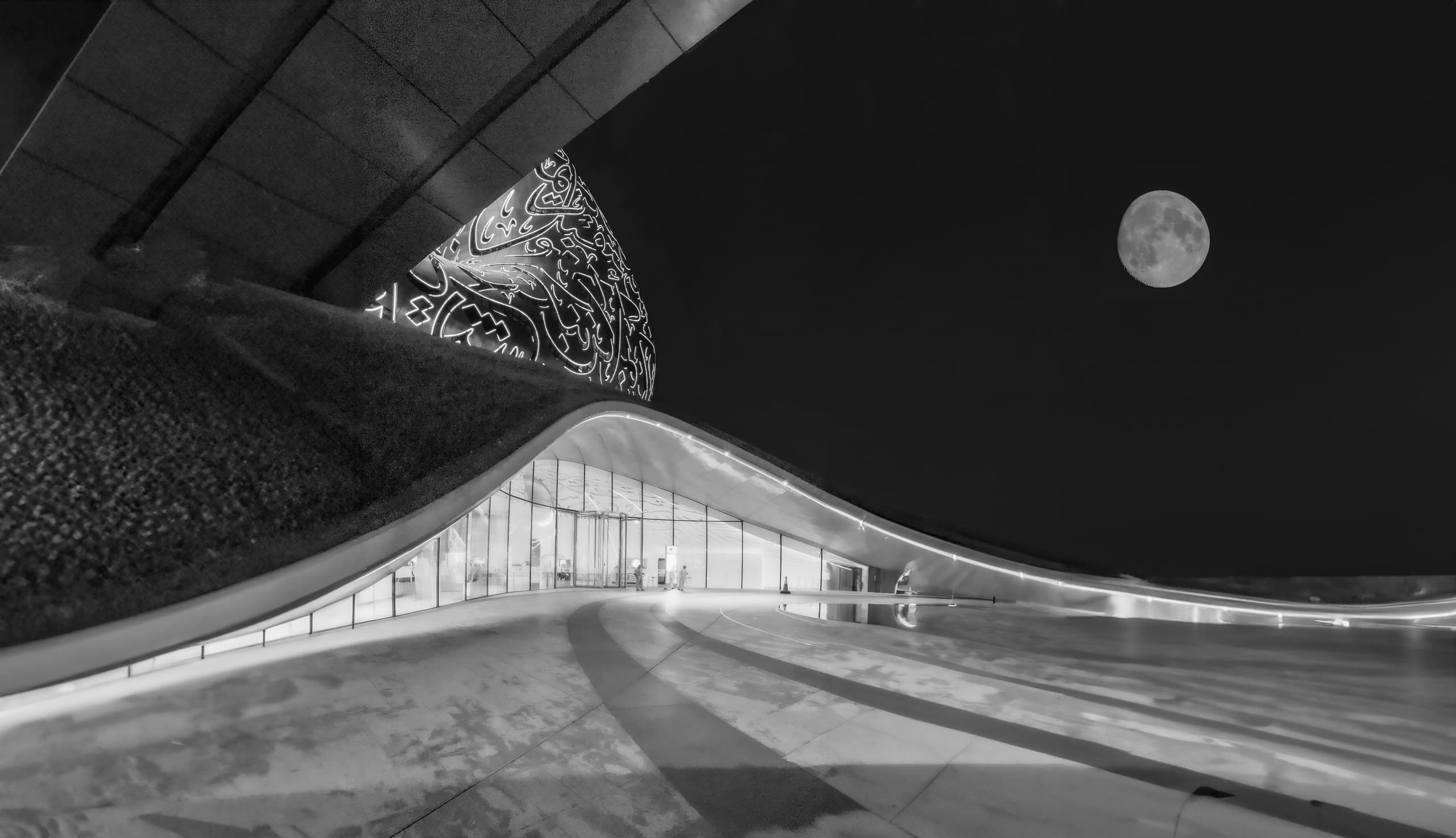 Black and white night photograph showing a curving modern building with illuminated arabic calligraphy windows and a bright full moon in the dark sky above.