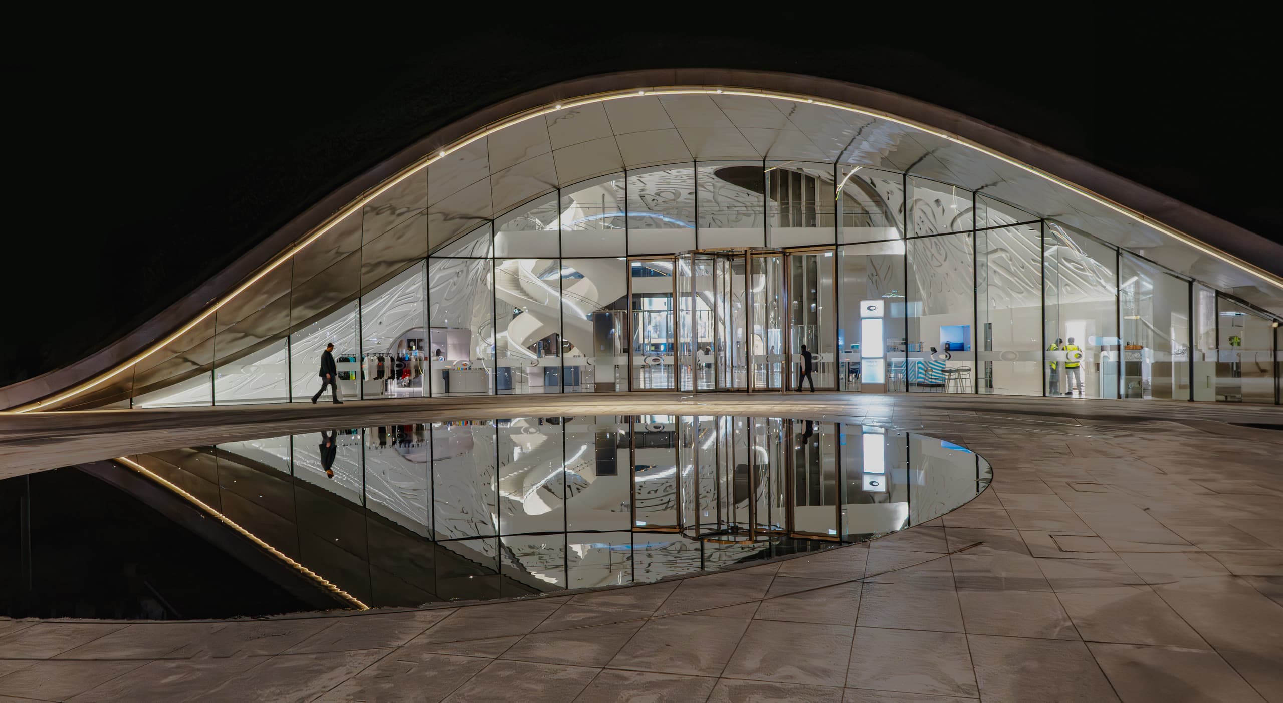 Night view of a low, wide modern building with a curved arch roof and glass facade, featuring a solitary person walking left and a puddle reflecting the illuminated entrance.
