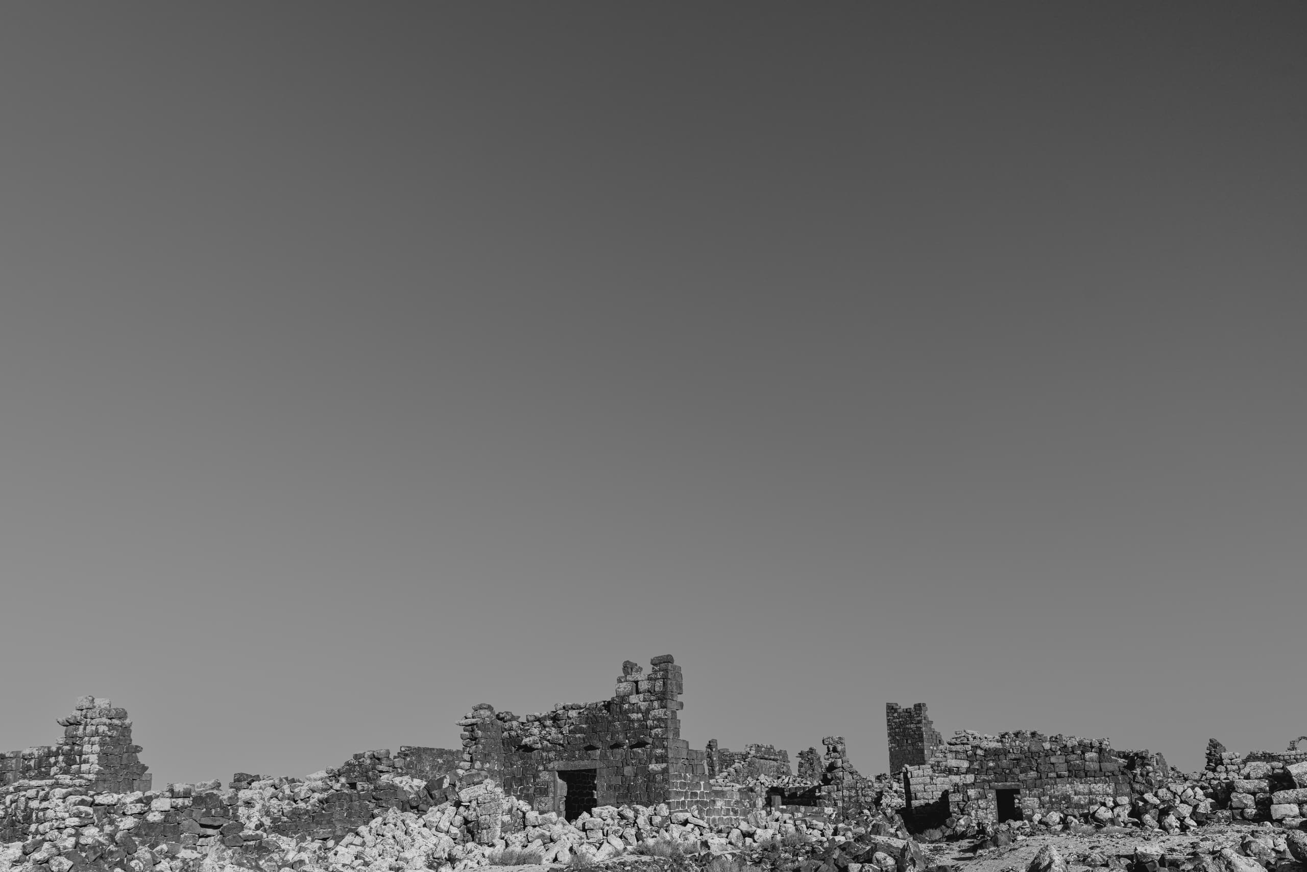 Low-angle black-and-white view of weathered ancient stone ruins and fragmented walls stretching horizontally across the lower third of the frame beneath a vast, unblemished sky.