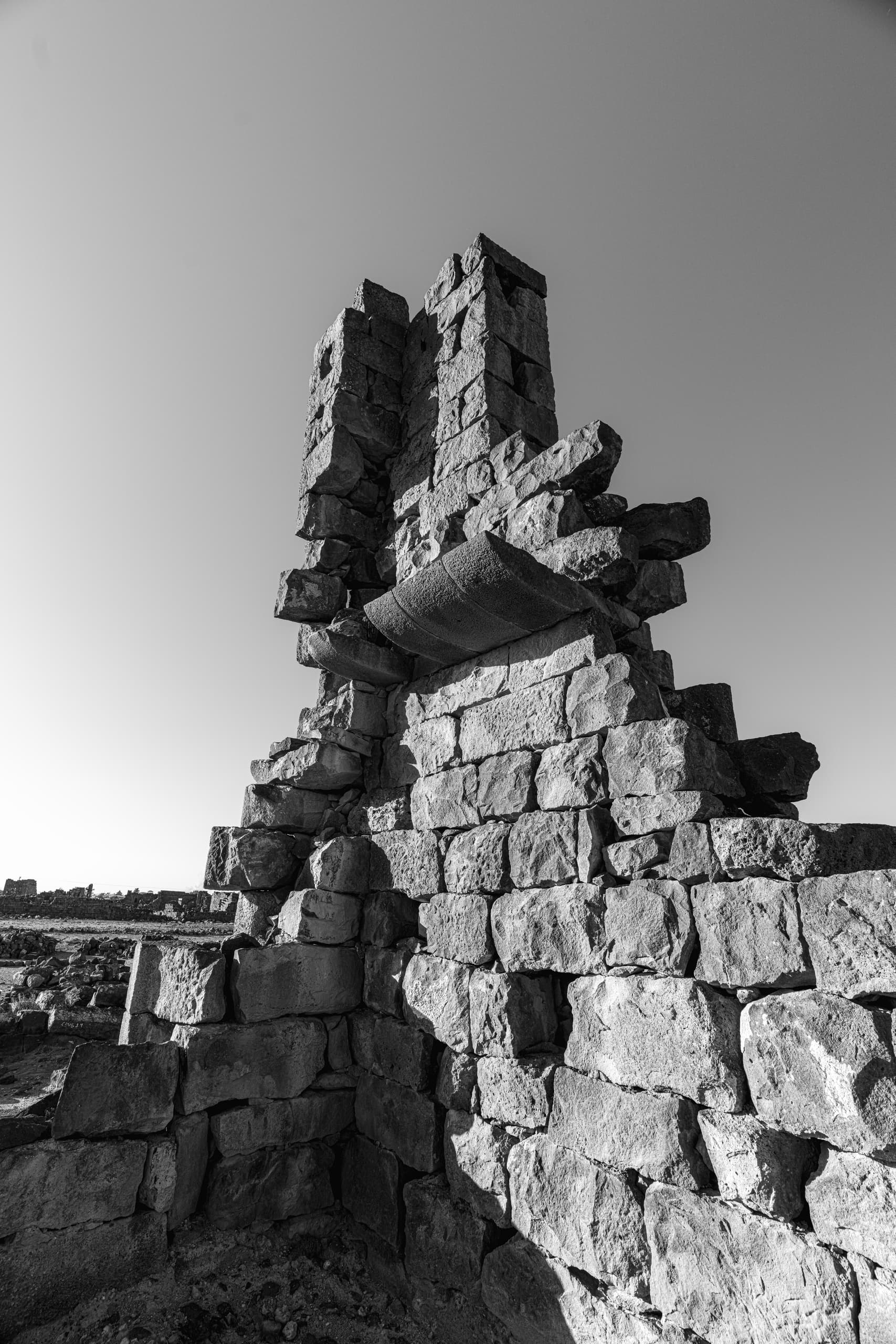 A low-angle monochrome photograph of ancient stone ruins featuring jagged blocks, weathered textures, and a clear sky.