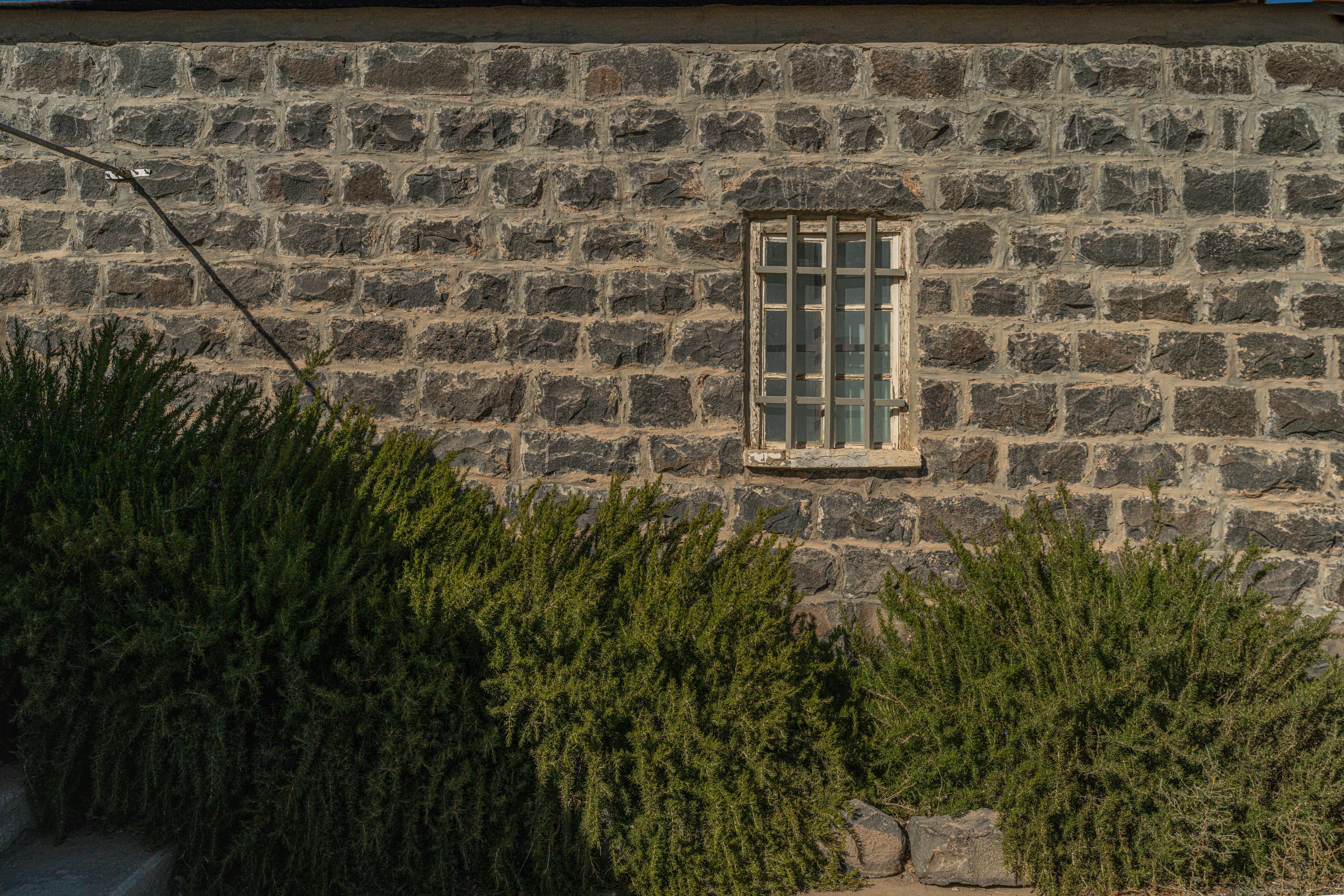 A weathered basalt stone wall featuring a small barred window and dense green bushes growing at its base, illuminated by directional light.