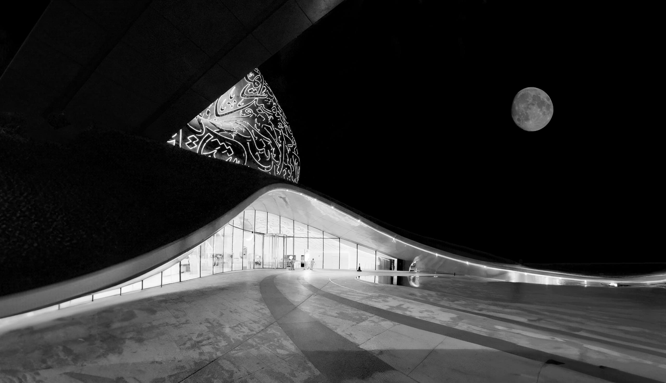 Black and white photograph showing a modern curved structure with illuminated Arabic calligraphy and a bright full moon visible in the black sky above the glass entrance.
