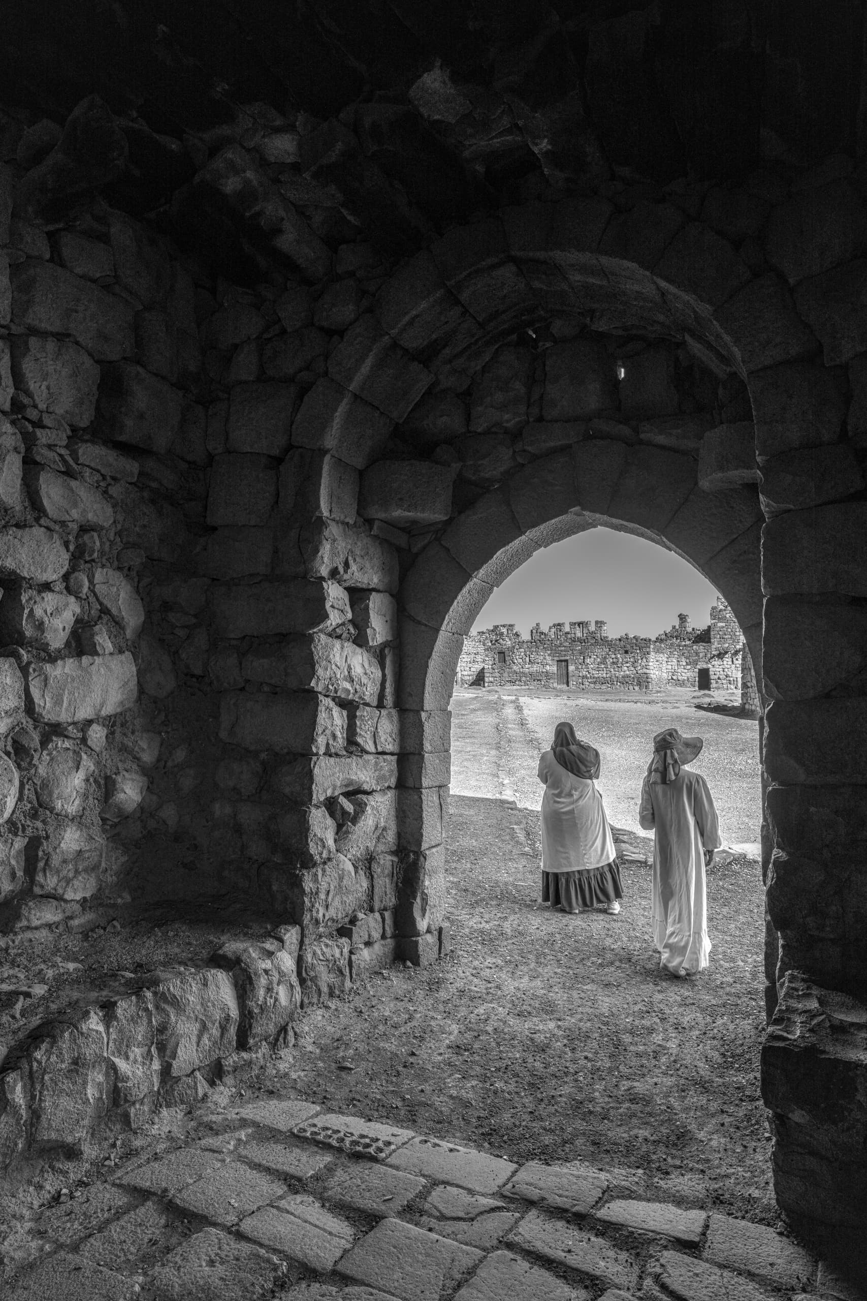 Monochrome interior view looking out through a rugged stone arched doorway into a bright open courtyard where two figures are seen walking away from the camera.