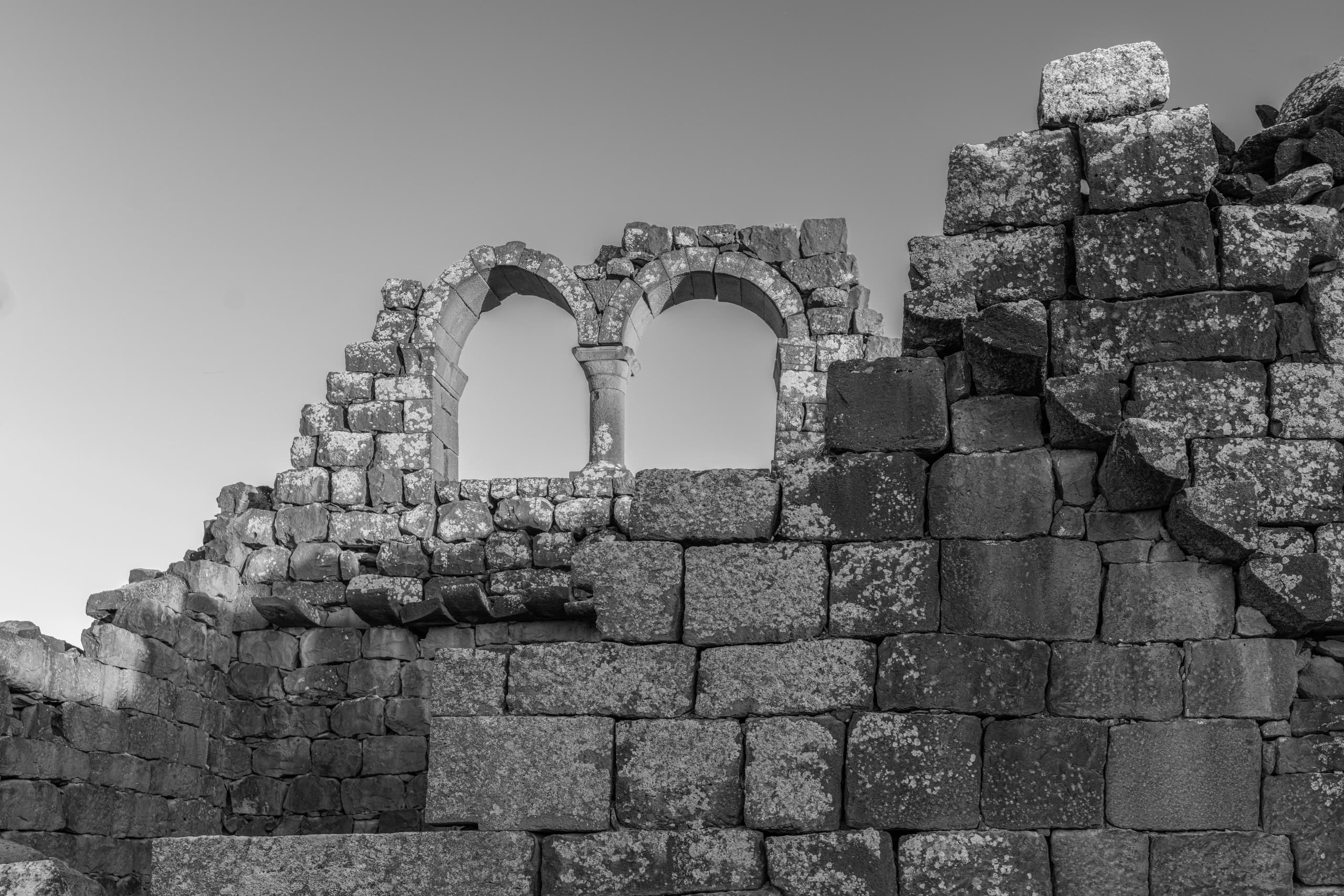Black and white photograph of ancient stone ruins, featuring a double arched window framed by weathered basalt blocks and a smooth gradient sky above.