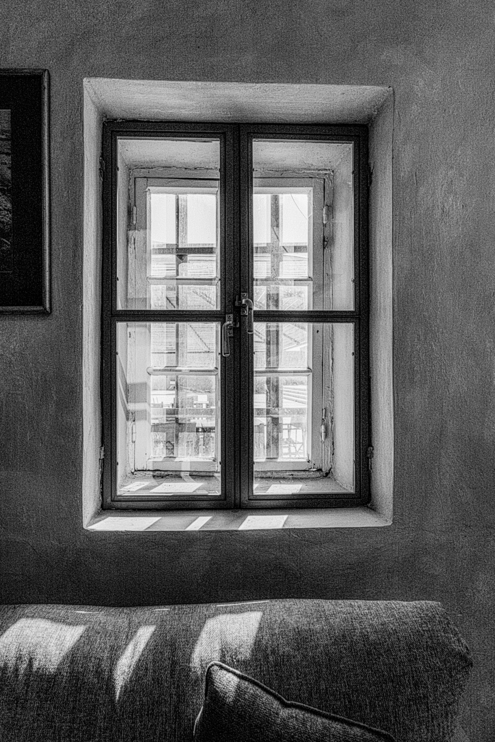 Monochrome interior view focusing on a multi-pane window recessed in a textured plaster wall, with angled sunbeams falling across a upholstered sofa back in the foreground.