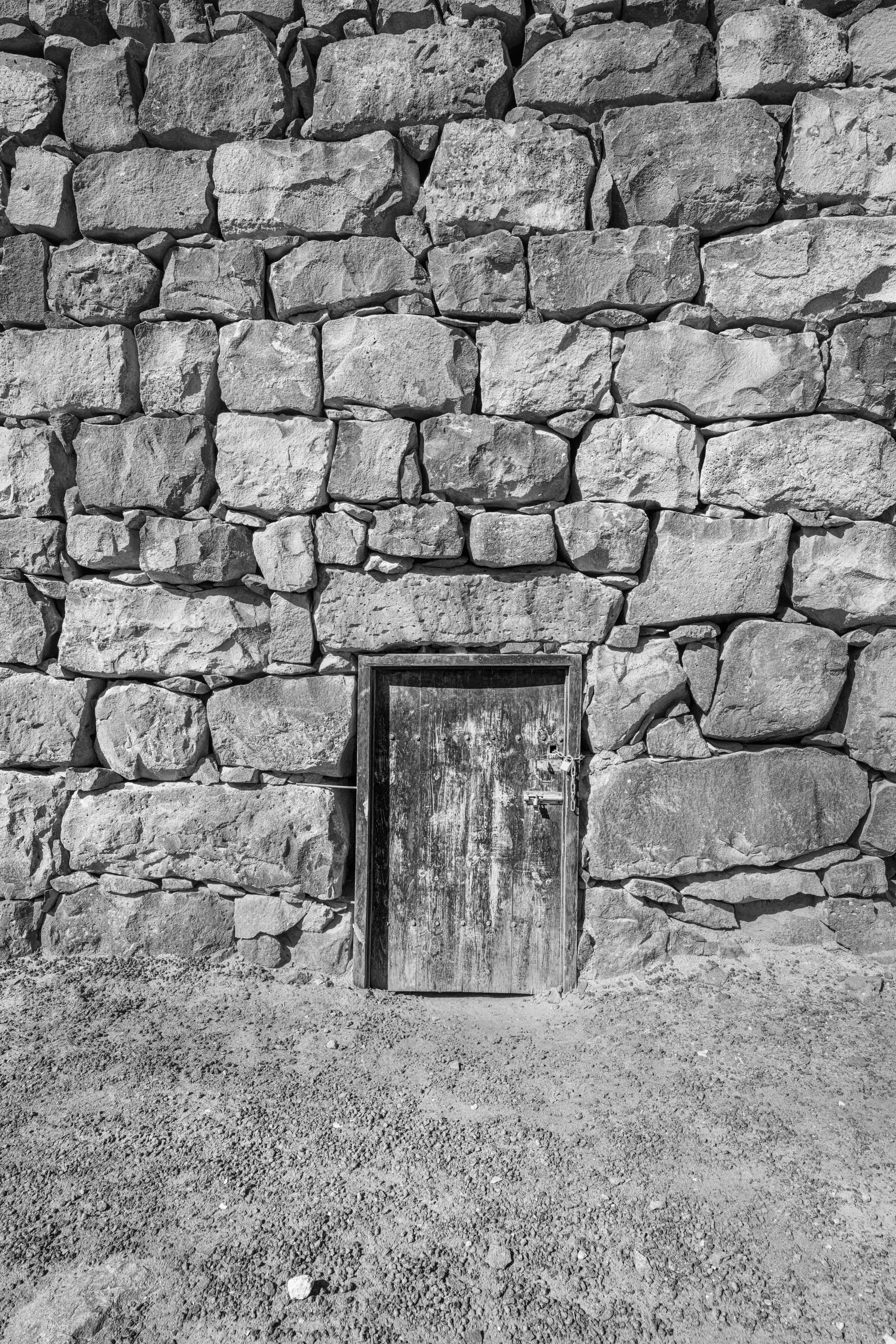 Black and white photograph of a small, aged wooden door integrated into a towering, rough-hewn stone wall above a gravel-covered ground.