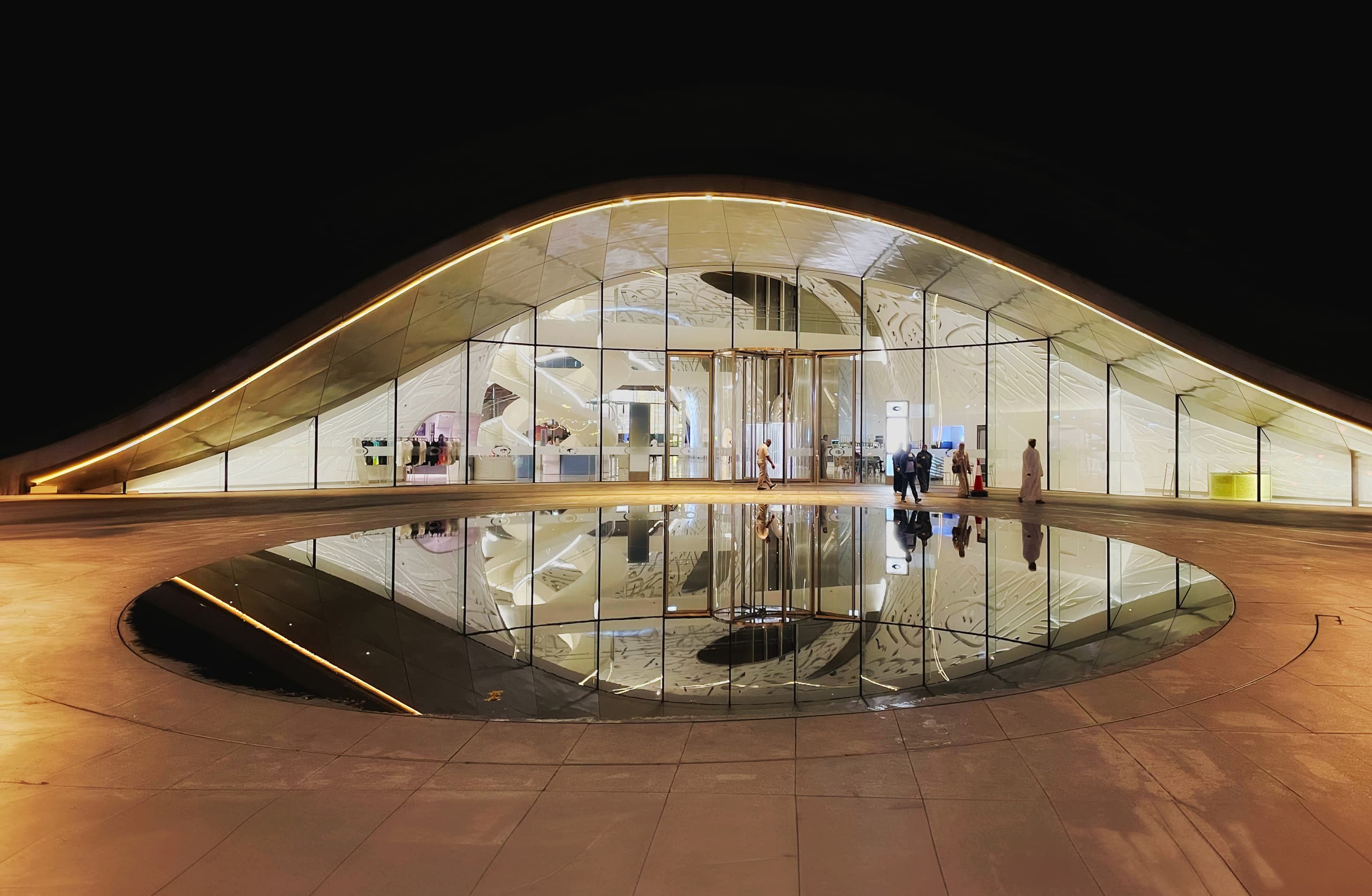 Night view of a low, curved modern building with a glazed entrance facade, reflected symmetrically in a circular pool in the foreground, with distant figures walking across the plaza under a black sky.