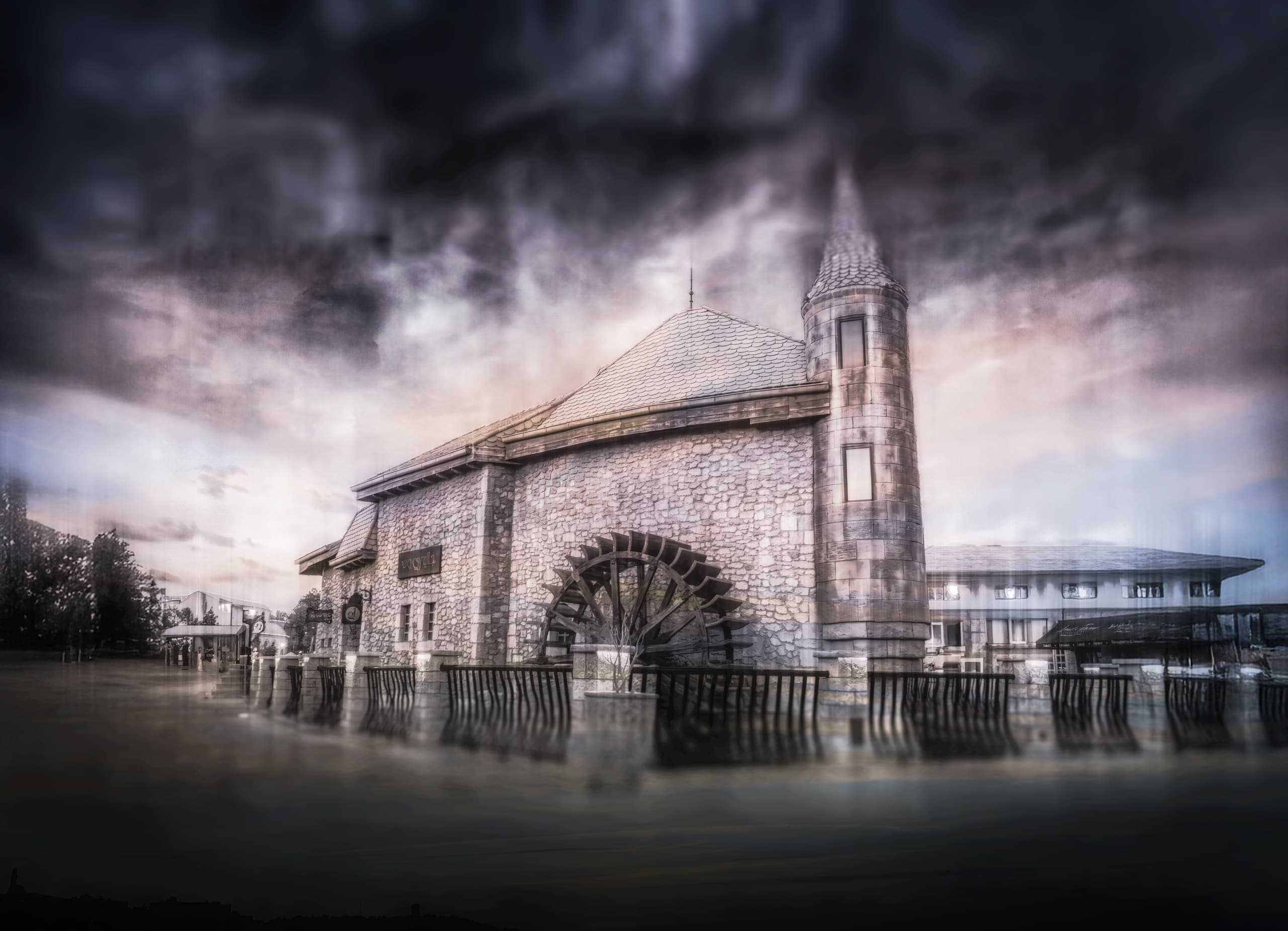 Exterior architectural view of a stone building with a large wooden water wheel and round tower under a dark, dramatic cloudy sky, with foreground motion blur.