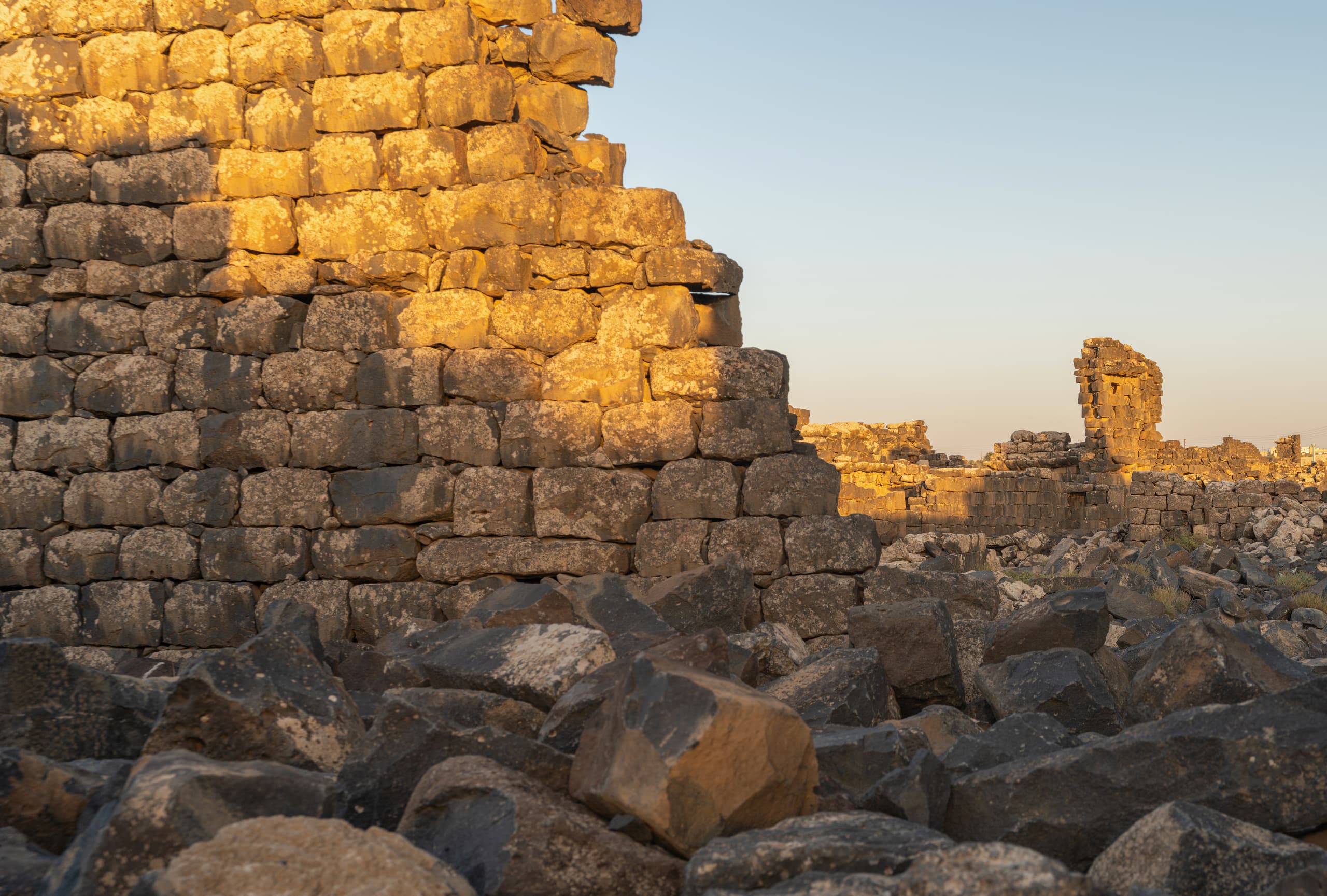 A low-angle view of a large, weathered stone wall partially illuminated by golden hour light, rising above a rough foreground of dark scattered basalt rocks and distant ruined foundations under a clear blue sky.