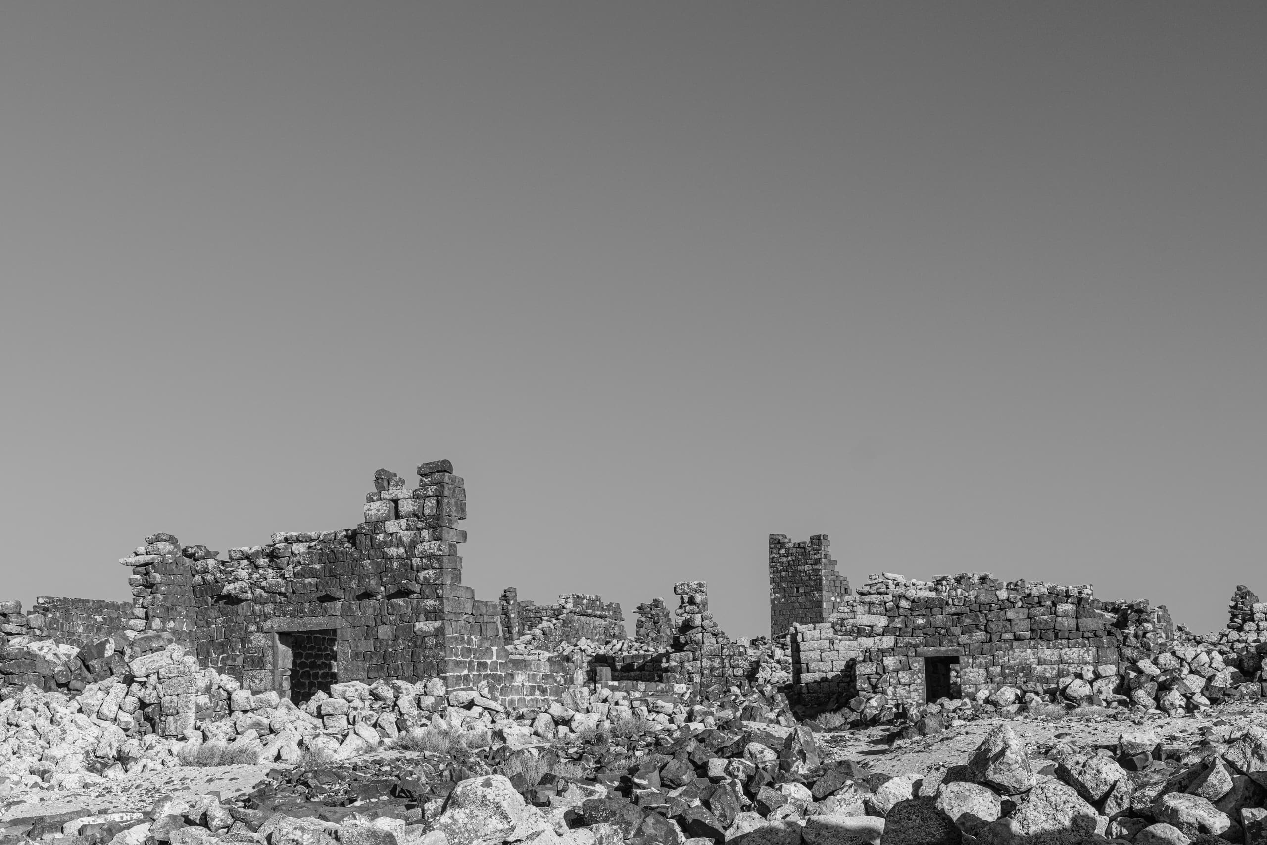 Black and white photograph showing crumbling ancient stone structures and scattered rubble across the ground under a clear, bright sky, with dark basalt masonry forming low walls and doorways.